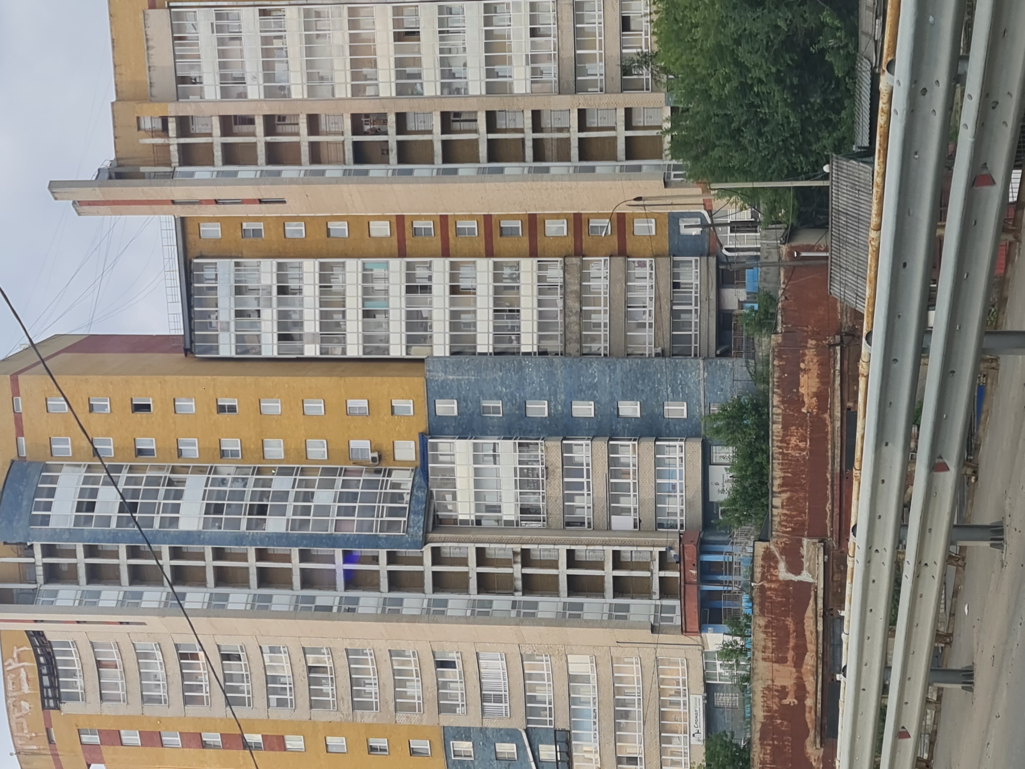 The image shows a residential building in the city of Irkutsk, Russia. The building is multi-story with a yellow facade and blue accents. It has numerous windows and balconies. In the foreground, there is a metal railing, likely part of a bridge or overpass, suggesting the photo was taken from a elevated position. Behind the railing, there is a rusty metal structure and some greenery. The sky is overcast. No people are visible in the image. The overall scene depicts an urban environment with residential architecture.
