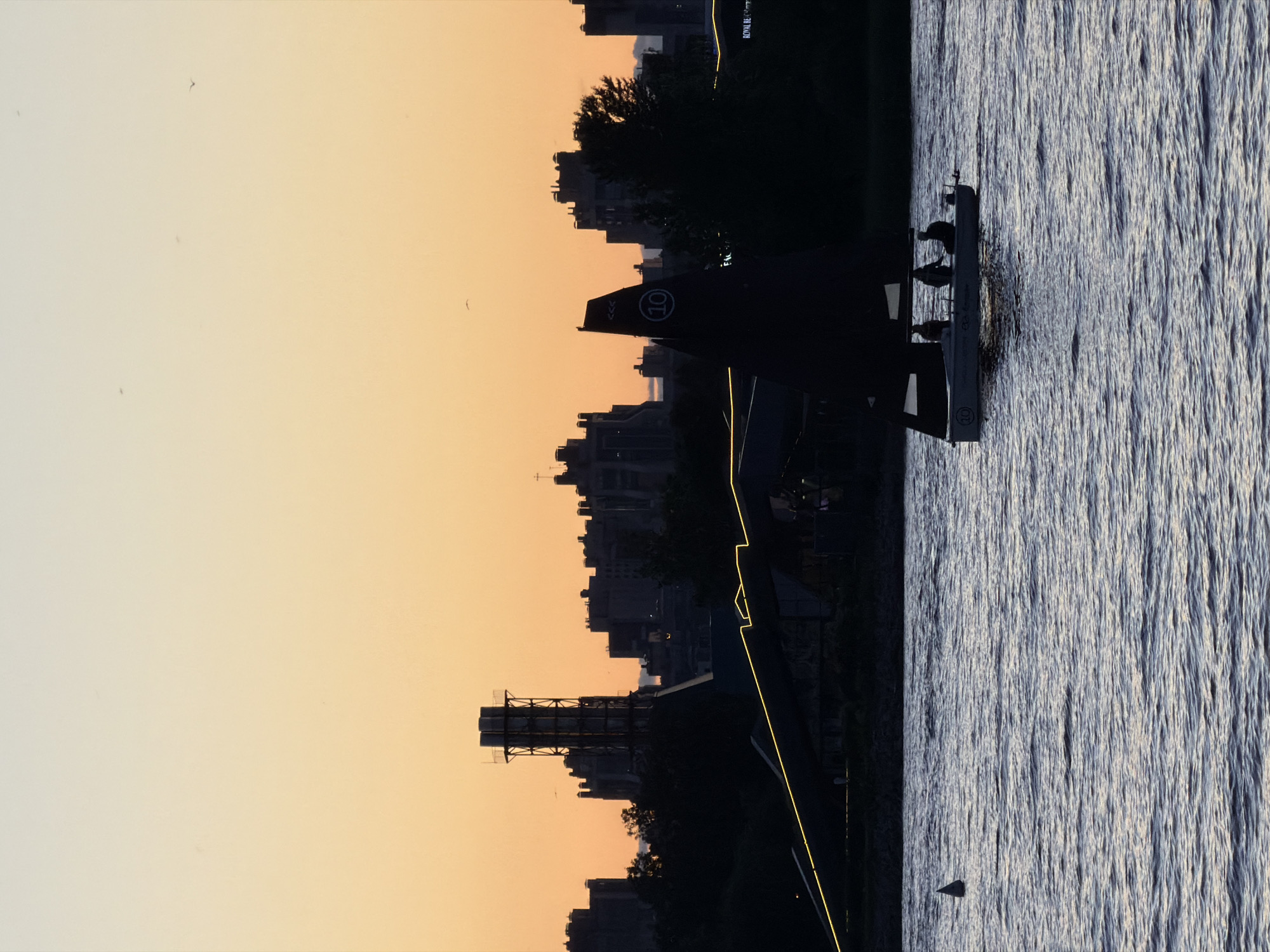 The image depicts a serene sunset over a body of water, likely the Neva River, in Saint Petersburg, Russia. Silhouetted against the orange and yellow sky are the distinctive outlines of the city's industrial and residential buildings, including a tall industrial tower. In the foreground, a small sailboat with a dark sail glides across the water, carrying a group of people whose figures are indistinct in the low light. The water's surface is rippled, reflecting the muted colors of the sky. A small buoy is visible to the left of the sailboat. The overall scene evokes a sense of tranquility and the end of a day in a bustling urban environment.