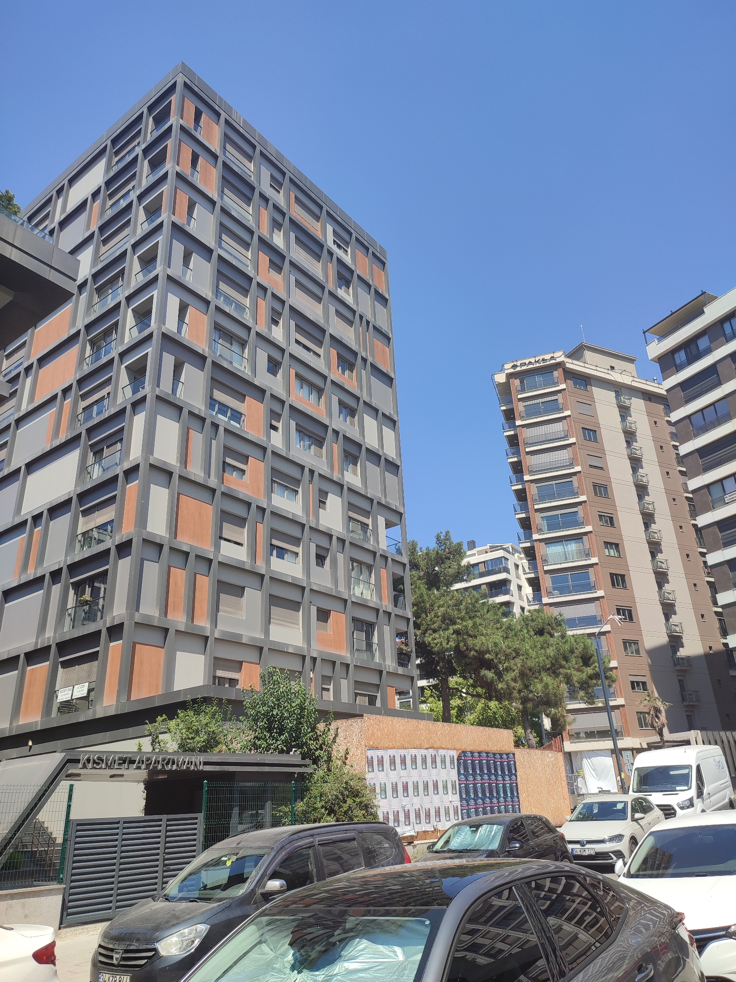 The image shows a street view in Kadıköy, Turkey, on a clear, sunny day. In the foreground, several cars are parked, including a dark-colored SUV, a white sedan, and the rear of a white car. Behind the cars, a low-rise building with a slatted metal fence and a sign reading "KISMET APARTMANI" is visible. Lush green trees are planted around this building.
Dominating the background are two modern apartment buildings. The building on the left is a tall, multi-story structure with a dark grey facade featuring a geometric pattern of orange-colored rectangular accents. Its exterior is characterized by recessed balconies and numerous windows. To the right of this building stands a slightly taller, slender apartment building with a beige and brown facade. This building also features multiple balconies and many windows, and a sign at the top reads "PAKLA". Further to the right, another modern apartment building with a white and grey striped facade and balconies can be seen. In between the buildings, there are more trees and glimpses of other structures. A white van is parked on the street. The sky is a clear, bright blue.