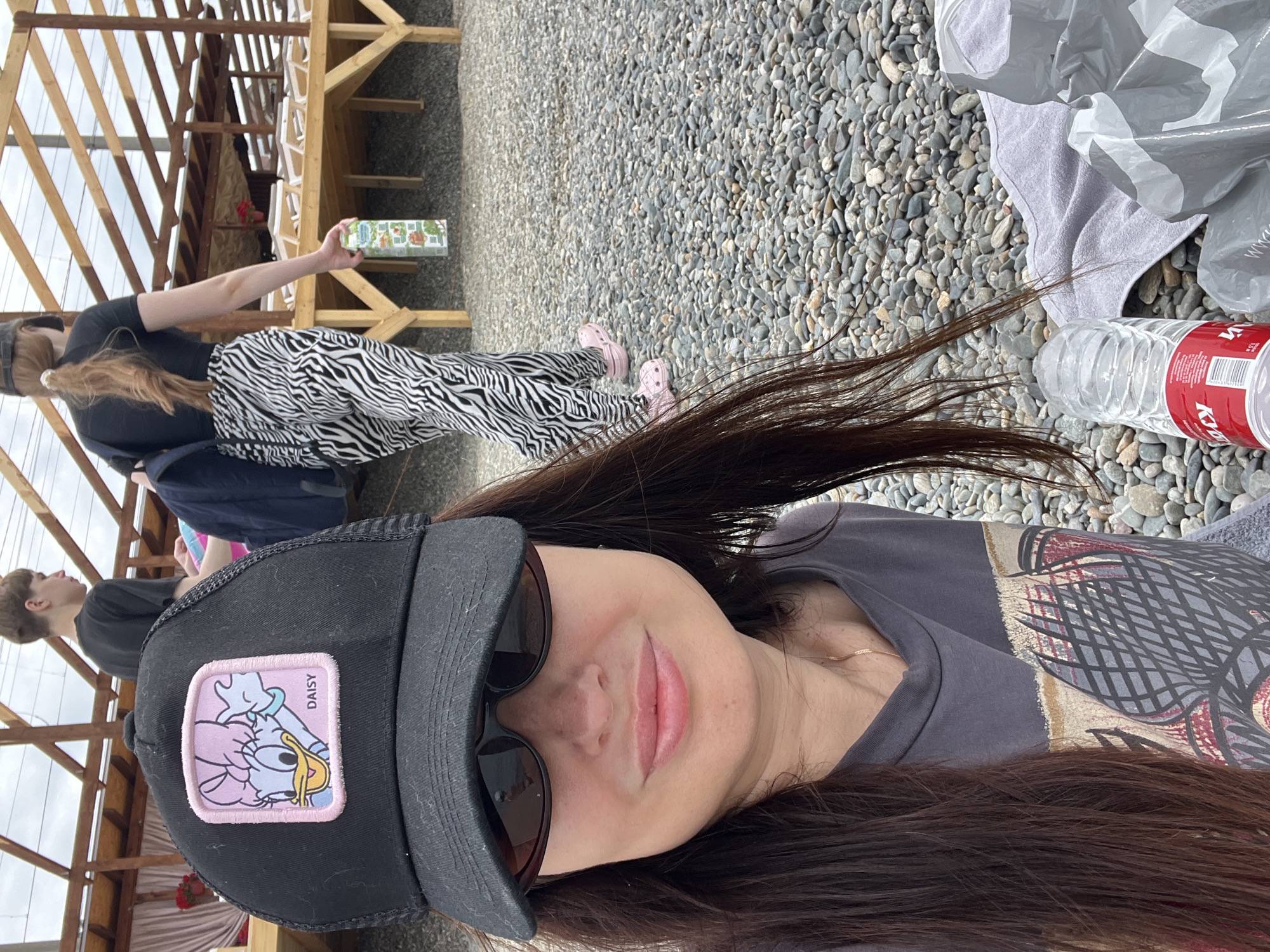 In an outdoor setting with a gravel ground and a wooden structure in the background, a woman wearing a black baseball cap with a Daisy Duck patch and sunglasses is in the foreground, taking a selfie. Her dark hair is long and extends past her shoulders. She is wearing a gray t-shirt with a graphic design. Behind her, a person with short hair is visible, and further back, another person wearing a black top and zebra-print pants is walking away from the camera, holding a juice box. The wooden structure appears to be a covered seating area with benches or lounge chairs. There is a water bottle and plastic bags on the ground near the woman.