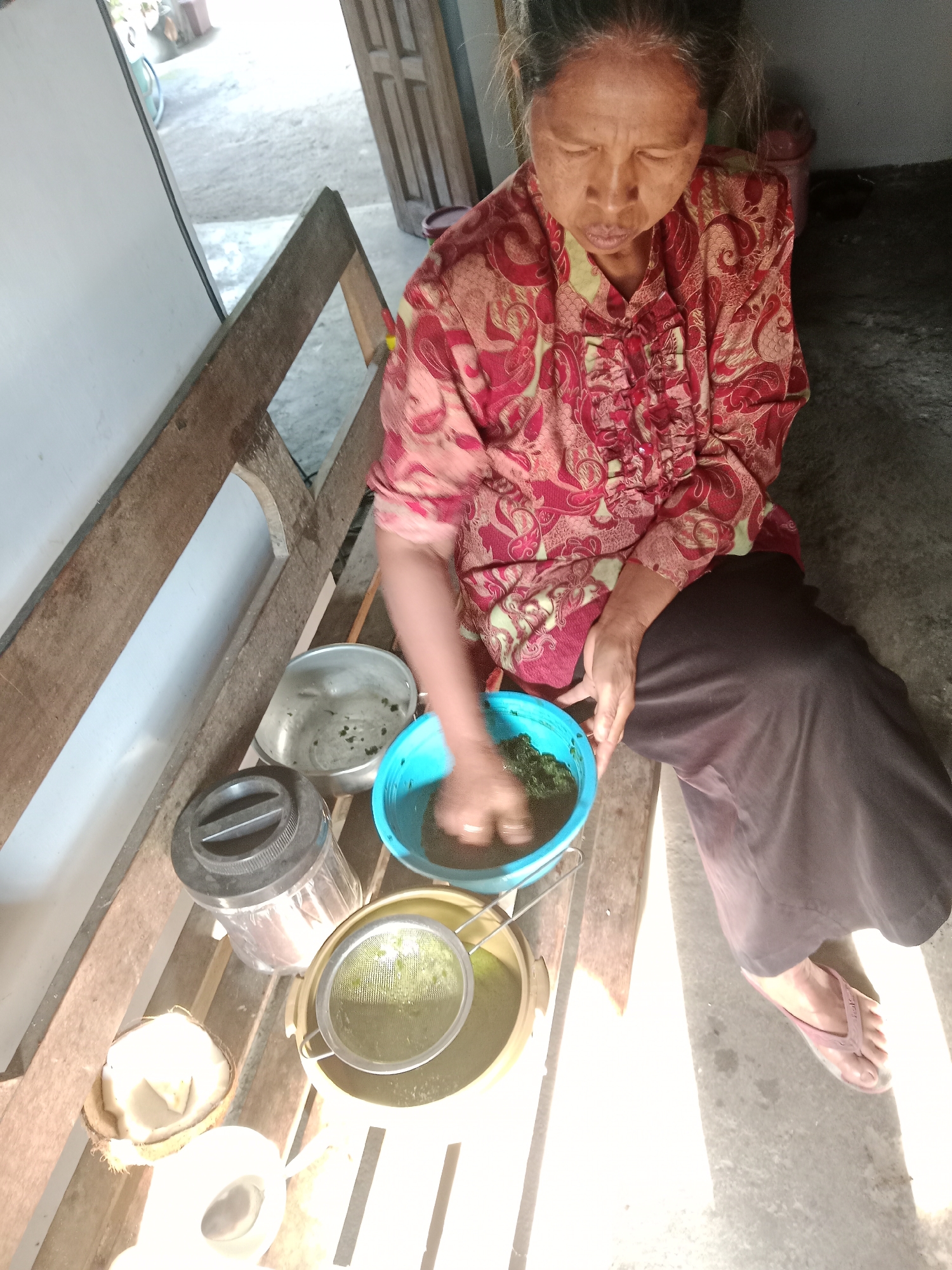 In the village of Sumberasri, Indonesia, an elderly woman with dark hair is seated on a rustic wooden bench. She is wearing a vibrantly patterned red and pink blouse with intricate detailing and a long, dark brown skirt. Her eyes are closed, and her head is tilted downwards as she focuses on a task. In front of her, a bright blue plastic basin holds green leafy material, which she is processing with her hands. Adjacent to the blue basin is a silver strainer placed over a yellow bowl. To her left, a clear glass jar with a black lid is visible, and further to the left, a halved coconut sits on the bench. The environment appears to be an outdoor or semi-outdoor space, with a concrete floor and wooden structures suggesting a simple dwelling or workspace. Sunlight streams in from the right, casting distinct shadows across the scene.
