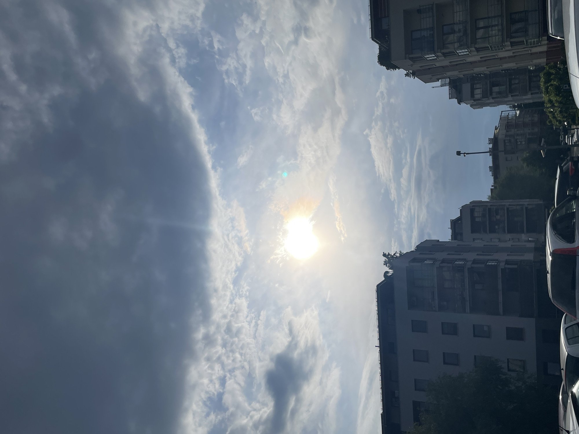 The image depicts a view of buildings and sky, with the sun shining brightly through scattered clouds. The buildings are multi-story apartment-style structures, and several cars are parked on the street alongside them. The image appears to be taken at street level, looking up, and is oriented vertically. The exact location and time are unknown, but the presence of buildings and cars suggests an urban environment.