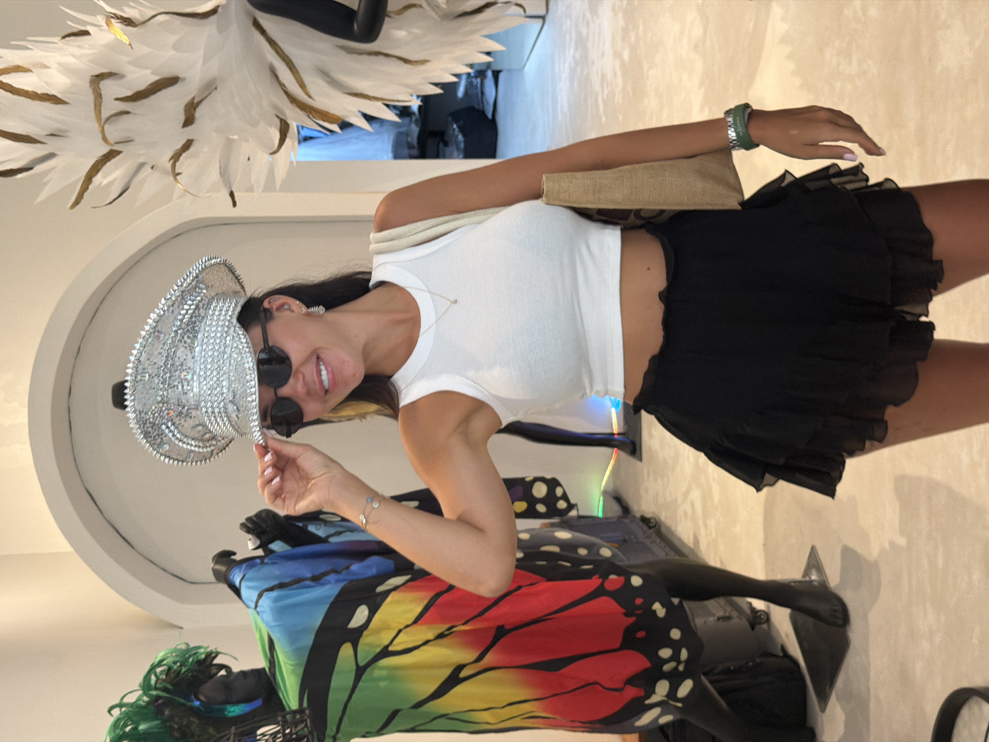 A woman wearing a white crop top, black ruffled skirt, sunglasses, and a bedazzled cap is posing for a photo in what appears to be an indoor retail or exhibition space. She is holding the brim of her cap with her left hand and smiling at the camera. To her left, a mannequin is dressed in a colorful butterfly-wing-inspired garment. To her right, a large, abstract white and gold feathery sculpture is visible. The floor is a light-colored, smooth surface. The background shows arched doorways and what seem to be other display elements. The scene is well-lit, with soft lighting. The woman's attire suggests a festive or avant-garde style. There are no visible indications that this location is in Ko Samui District, Thailand.