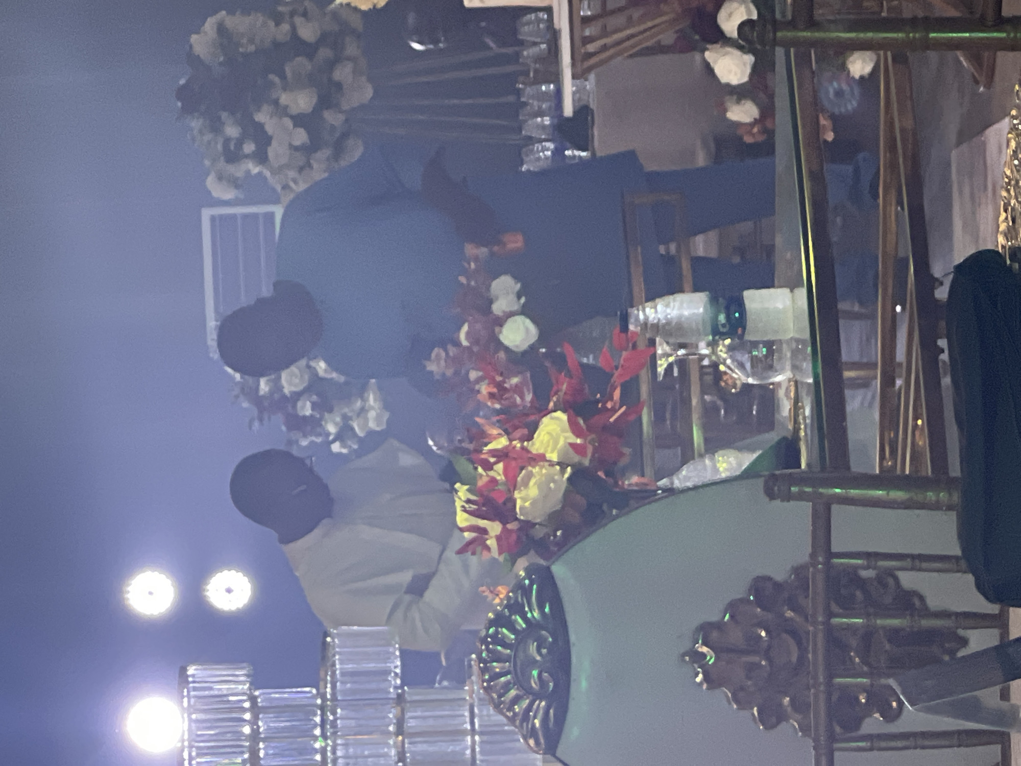 The image depicts a wedding reception setting in Abuja, Nigeria. Two men in traditional Nigerian attire (one in white and the other in blue) are arranging floral centerpieces on a table. The table is decorated with ornate gilded details. The background features crystal glassware, and large floral arrangements. Lighting fixtures are visible in the upper and lower portions of the frame. The overall setting suggests a formal indoor event space.