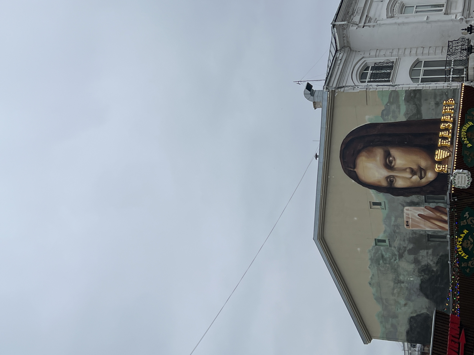 The image shows the side of a building in Kazan, Russia. The building has a large mural of a woman in a headscarf taking a selfie with a mobile phone. Above the mural is a white building with arched windows. To the right of the mural is a sign with neon lights that reads "ТАБАТ Аш". The sky is overcast.
