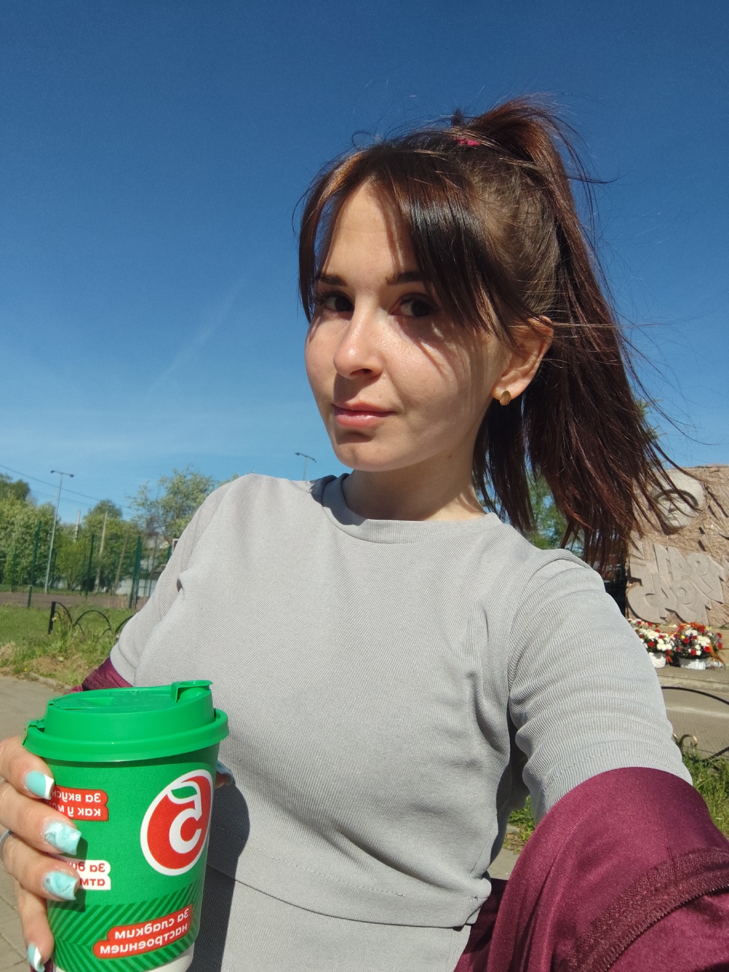 A woman is taking a selfie outdoors on a sunny day. She is holding a green coffee cup with Russian text and the logo of the brand "Пятёрочка." The woman has brown hair in a high ponytail with bangs, wearing a gray long-sleeved shirt, and a maroon jacket sleeve is visible. Her fingernails are painted light blue and white.
The background consists of a clear blue sky, green trees, grass, and a memorial or monument adorned with red and white flowers. The location is Фоки, Russia, based on the text on the coffee cup.
