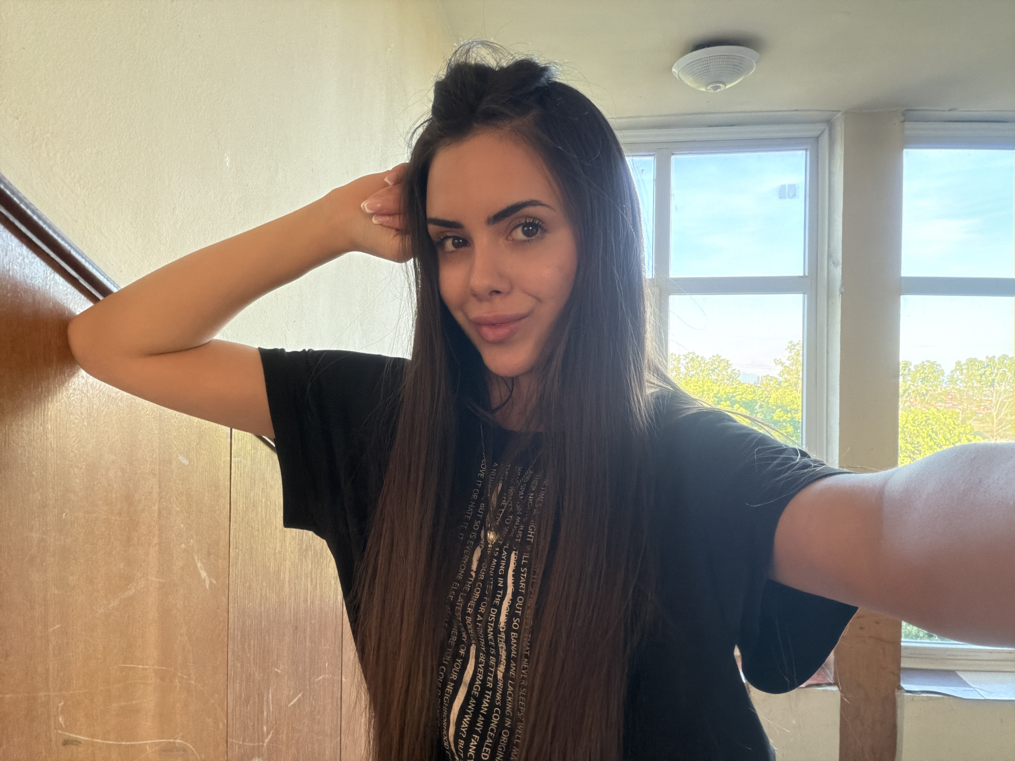A woman with long, dark hair is taking a selfie indoors, leaning against a wooden wall. She is wearing a black t-shirt with white text printed on it. The background shows a window with a view of trees and sky. A light fixture is visible on the ceiling. There is nothing in the image to suggest the location is Sofia, Bulgaria.