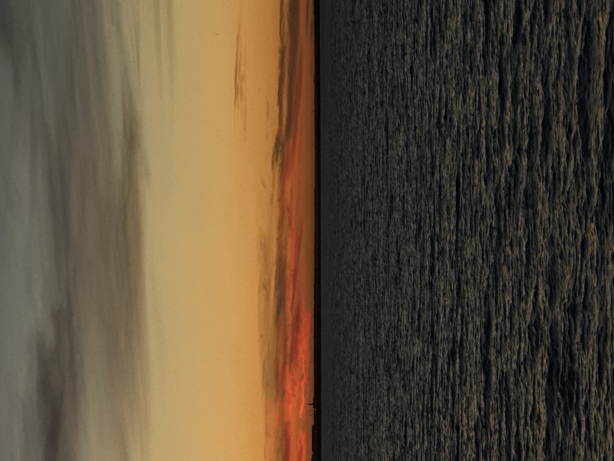 The image depicts a sunset over a body of water. The sky transitions from a dark gray at the top to a vibrant orange and red near the horizon. A thin, dark line representing land is visible in the distance, with faint structures that could be buildings or industrial elements. The water is dark and choppy, reflecting the subdued colors of the sky. The scene is serene and devoid of any human presence or activity. The location is specified as Kokshetau, Kazakhstan, suggesting a sunset over a lake or river within or near the city.