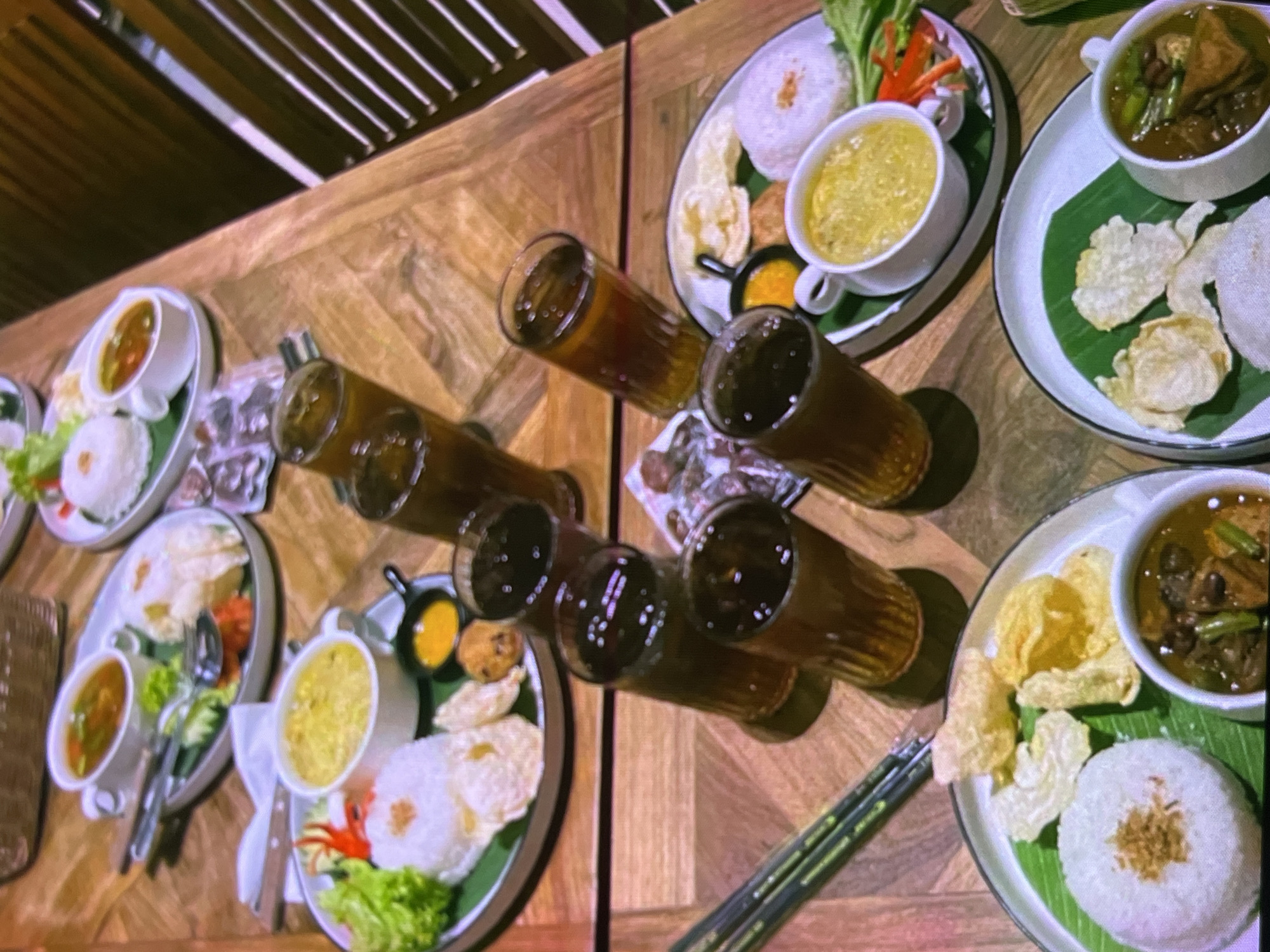 The image depicts a dimly lit, close-up overhead view of a wooden table set with several Indonesian meals and drinks, likely in a restaurant in Cadek, Indonesia. The meals consist of white rice, a side soup or curry in a white bowl, crackers, and some other accompaniments like fried shallots or garlic. There are also several glasses filled with a dark liquid, presumably iced tea or coffee. The setting appears to be a casual dining establishment, with the warm lighting contributing to a cozy atmosphere. No people are visible in the frame, and the focus is entirely on the food and drinks. The table's wood grain is prominent, suggesting a natural material used in the restaurant's decor.