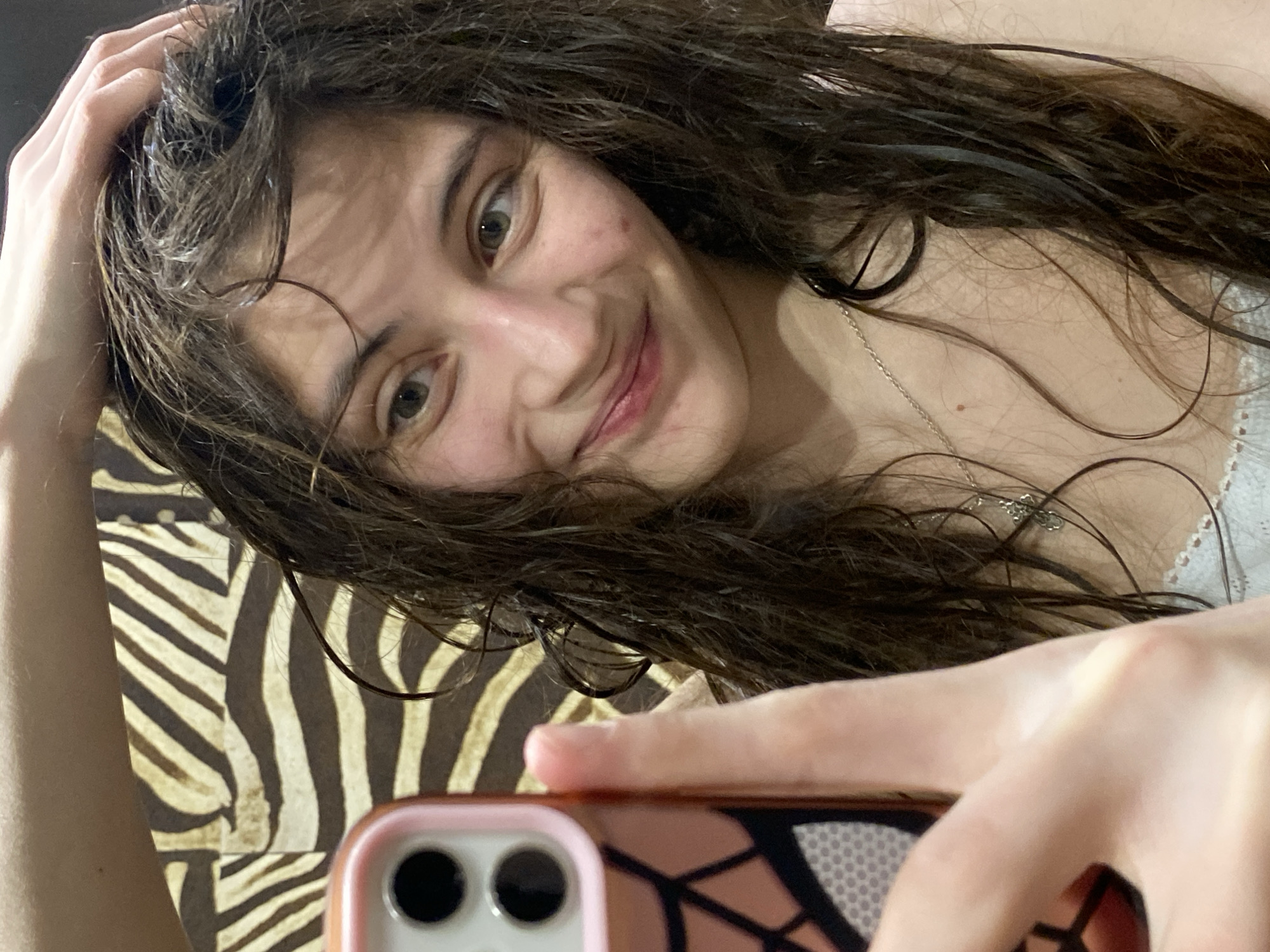 A young woman with wet, dark brown hair is taking a selfie. She has fair skin, light eyes, and is smiling. She is wearing a delicate silver necklace and a white lace top. Her left hand is touching her hair, and her right hand is holding a smartphone with a pink case featuring a geometric pattern. The background is a zebra-striped surface. There is no location information in the image, so it is impossible to determine if it was taken in Votkinsk, Russia.