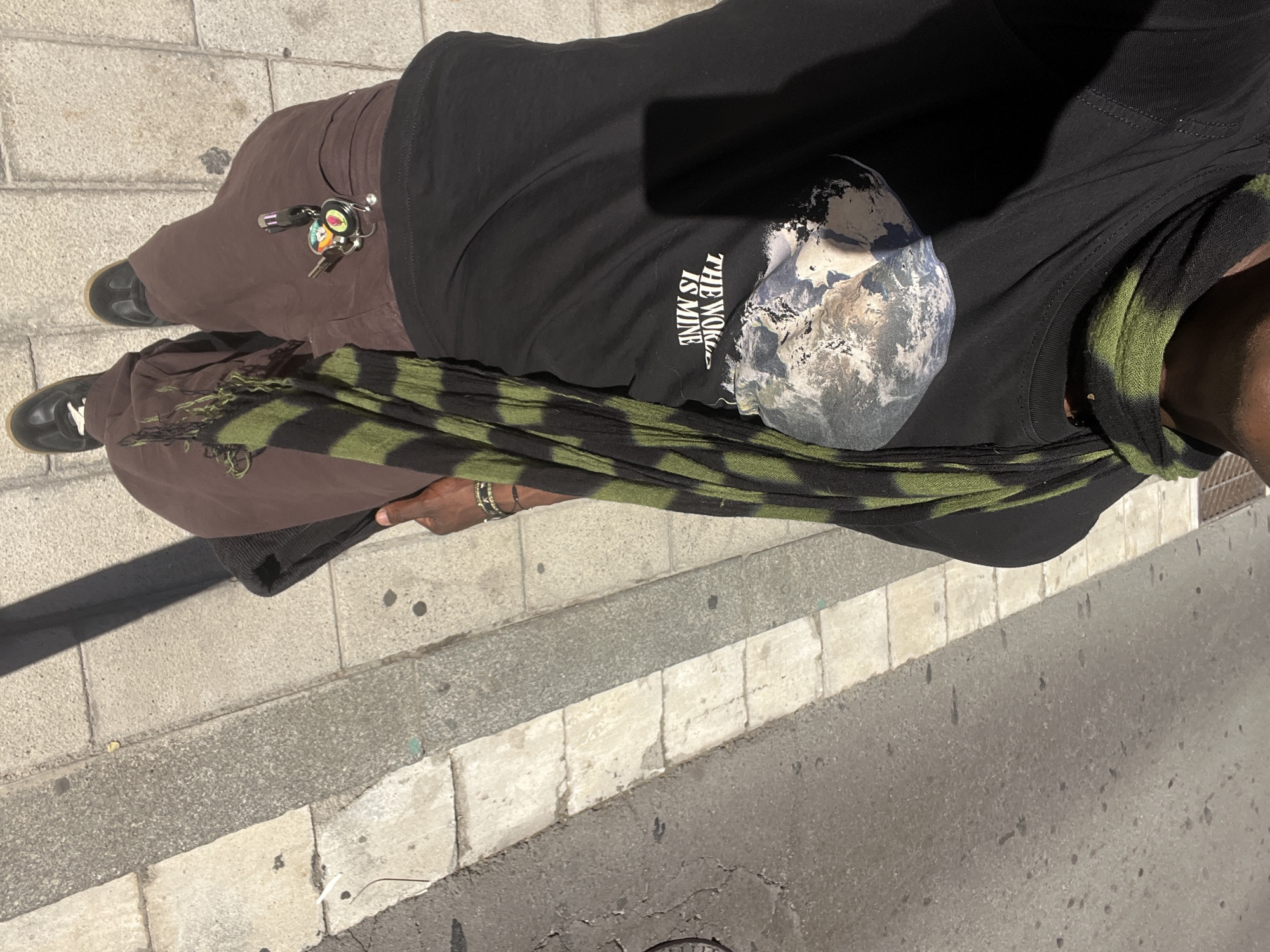 The image provides an overhead view of a person standing on a paved sidewalk next to a street, bathed in bright sunlight. The scene is consistent with an urban setting, such as Barcelona.
The person is wearing loose, brown pants and black shoes with light-colored soles. Attached to the pants is a keyring laden with several keys, a small flashlight, and at least two colorful charms—one resembling a cartoonish face and another an abstract shape. A distinctive green and black striped or camouflage-patterned scarf, featuring frayed edges, is draped around the person's upper body.
A black t-shirt is visible, displaying a graphic of the Earth alongside the prominent white text "THE WORLD IS MINE". On the visible wrist, the person wears multiple metallic bangles or bracelets.
The ground beneath is composed of light-toned, square-patterned sidewalk slabs adjacent to a darker asphalt street, clearly separated by a concrete curb. Long, distinct shadows indicate a sunny daytime. The image primarily captures the person's attire and immediate environment, with no discernible activity beyond standing or movement, and no visible facial expressions.