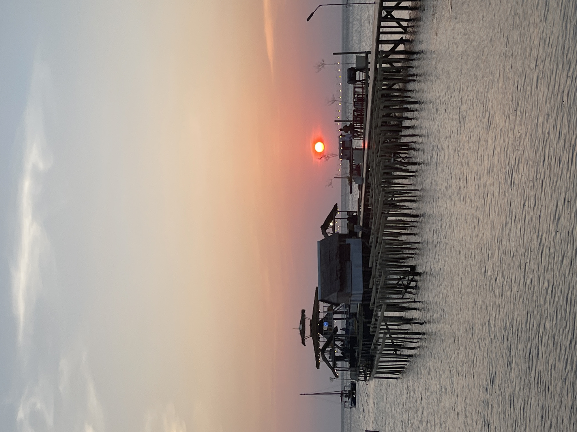 The image depicts a serene sunset over the ocean in Karimunjawa, Indonesia. The sky displays a gradient of soft pinks and oranges, with a prominent, bright orange sun nearing the horizon. Several wooden piers extend into the water. On the main pier, there are structures resembling huts or small buildings with thatched roofs, and some people are visible, seemingly relaxing or dining. A sailboat is docked to the left of the frame, and string lights are visible on a distant pier, suggesting a festive atmosphere. The water is calm, reflecting the colors of the sunset. The scene captures a peaceful evening ambiance in this Indonesian coastal town.