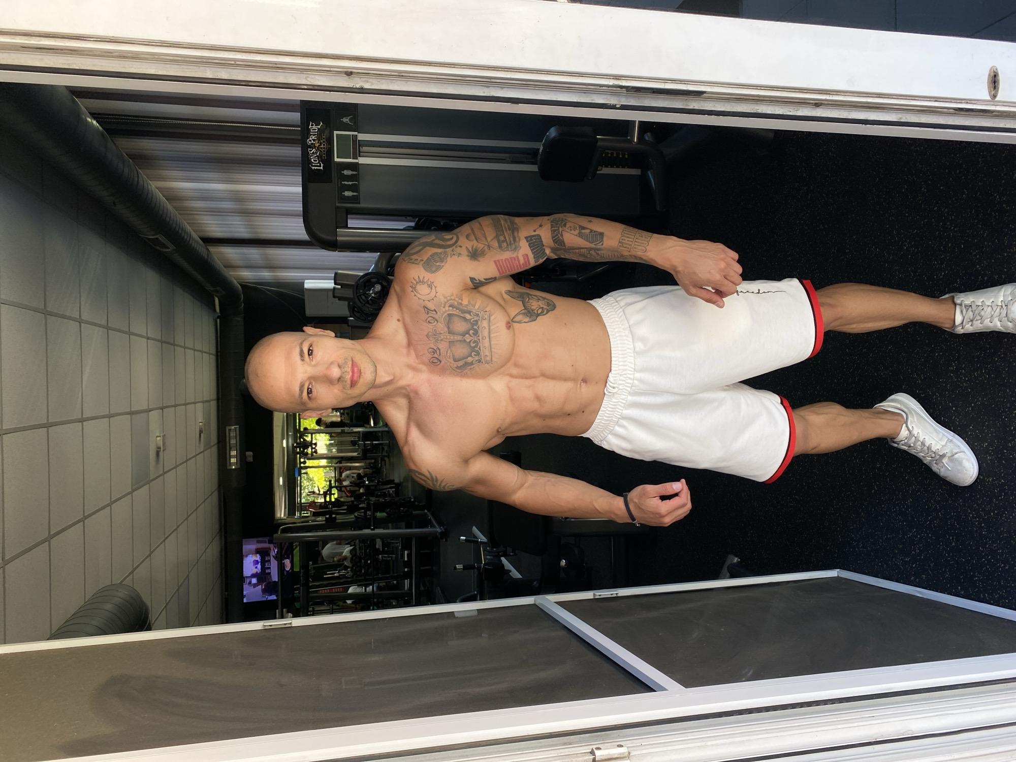 A muscular, bald man with numerous tattoos stands in a gym doorway in Pleven, Bulgaria. He is wearing white athletic shorts with red trim and silver sneakers. The gym is equipped with various weight machines and exercise equipment, with people visible in the background engaged in workouts. The lighting suggests it is daytime.