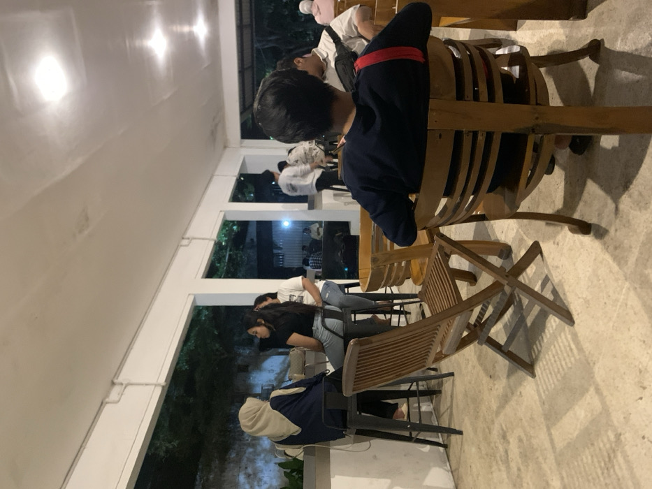 The image shows a scene inside an open-air cafe or restaurant in Probolinggo, Indonesia, located at coordinates -7.751 latitude and 113.213 longitude. There are four people seated at tables, with a fifth person standing nearby. The cafe is mostly empty, with wooden chairs and tables arranged throughout the space. The walls are white and there are large windows that offer views of the surrounding area. The light is dim, with light coming from ceiling lights. One person is sitting with their back to the camera, facing an unseen person behind them. Another person is seated on a wooden chair with their legs crossed, facing the left of the image. One person is seated on a stool, with their legs crossed and facing the right. Another person is seated on a stool with their legs crossed, facing the right, and appears to be conversing with the person sitting on the wooden chair. One person is standing behind the seated individuals, facing the left of the image. All of the people in the image are wearing clothing that covers their arms and legs. The cafe is set in a peaceful location, with the sounds of nature faintly audible.