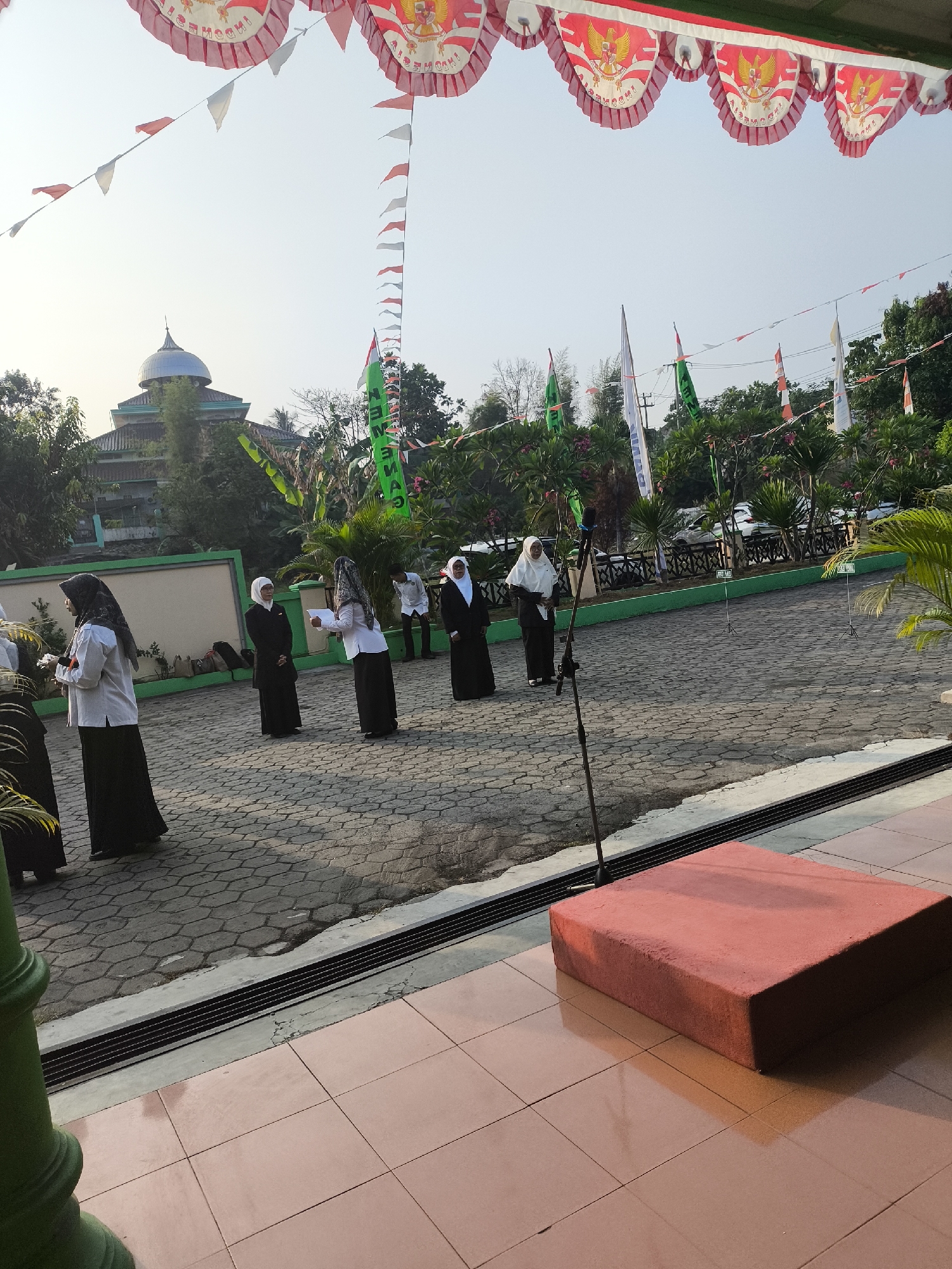 In Cilegon, Indonesia, an outdoor event is underway, characterized by festive decorations. A line of individuals, predominantly female and dressed in dark skirts and white tops with headscarves, stand in formation on a paved courtyard. One individual on the right holds a piece of paper, appearing to read aloud. Behind them, a building with a green roof and a dome is visible, surrounded by trees. The scene is further enhanced by numerous Indonesian flags and pennants strung across the area, some featuring the Garuda Pancasila emblem and the word "INDONESIA." A microphone stand is positioned in the foreground, and a red, cushioned seating platform is situated to the right, casting a shadow. The time of day appears to be morning or late afternoon, given the angle of the sunlight.