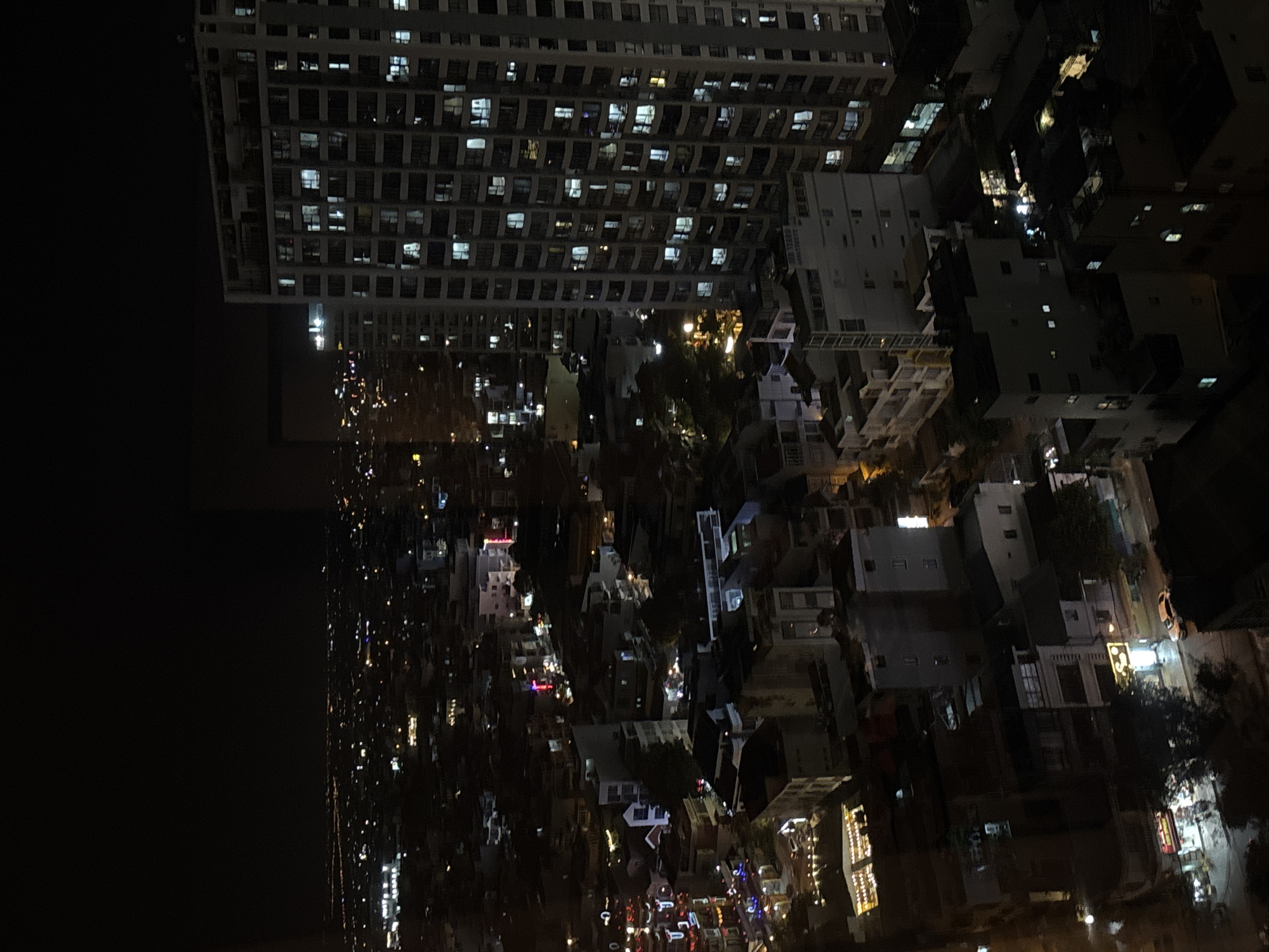 The image is a nighttime aerial view of a densely populated urban area, likely in Nha Trang Ward, Vietnam. The scene is dominated by buildings of varying heights, with a prominent, tall, modern apartment building on the right side of the frame. Many windows in this building are illuminated, suggesting occupancy. Below and to the left of the tall building, there are numerous smaller buildings, some appearing to be houses or low-rise apartments, with scattered lights indicating activity. In the distance, the lights of the city stretch towards the horizon, blurring into the darkness. Some buildings are decorated with colorful lights, possibly indicating commercial establishments or festive decorations. The overall impression is of a bustling city at night, with a mix of modern and older architecture. There are no visible people or specific actions depicted.