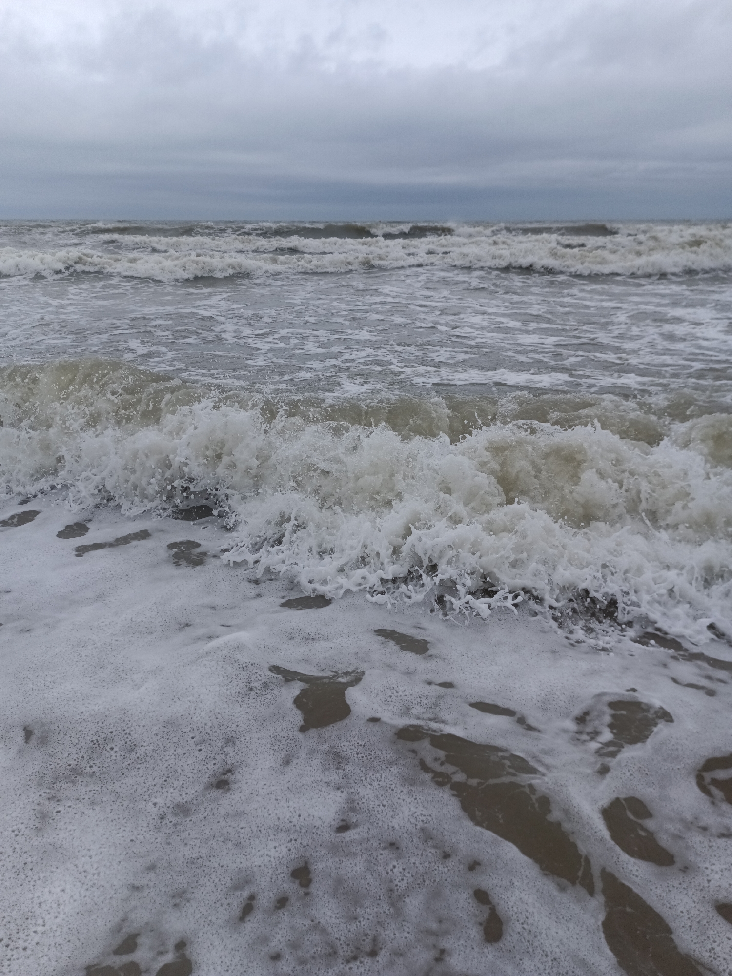 A wide-angle, eye-level shot captures a turbulent sea under a heavily overcast sky in Houlgate, France. The foreground is dominated by frothy white seafoam receding from the shore, revealing patches of wet sand. Beyond the foam, the ocean is agitated, with multiple rows of rough, grey waves breaking and churning. The horizon line is indistinct due to the low-lying grey clouds that completely obscure the sky. There are no people or man-made objects visible in the scene.