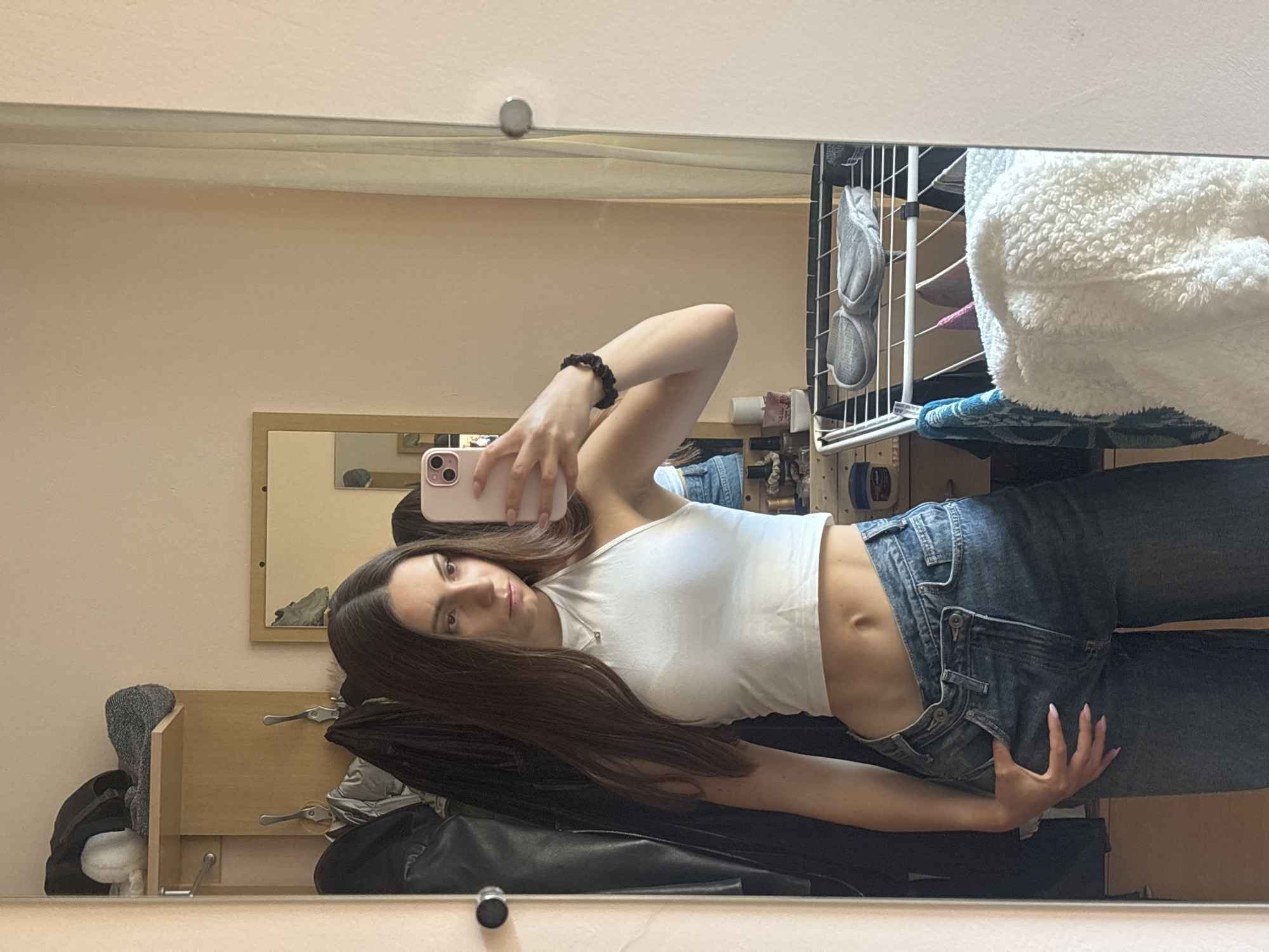A young woman with long, dark hair is taking a mirror selfie in a room. She is wearing a white halter crop top and dark wash, low-rise jeans. Her hand is resting on her hip, and she is looking at her reflection. In the background, there is a wooden closet with coats hanging on it, a drying rack with clothes and shoes, and a shelf with various items. The room appears to be domestic.