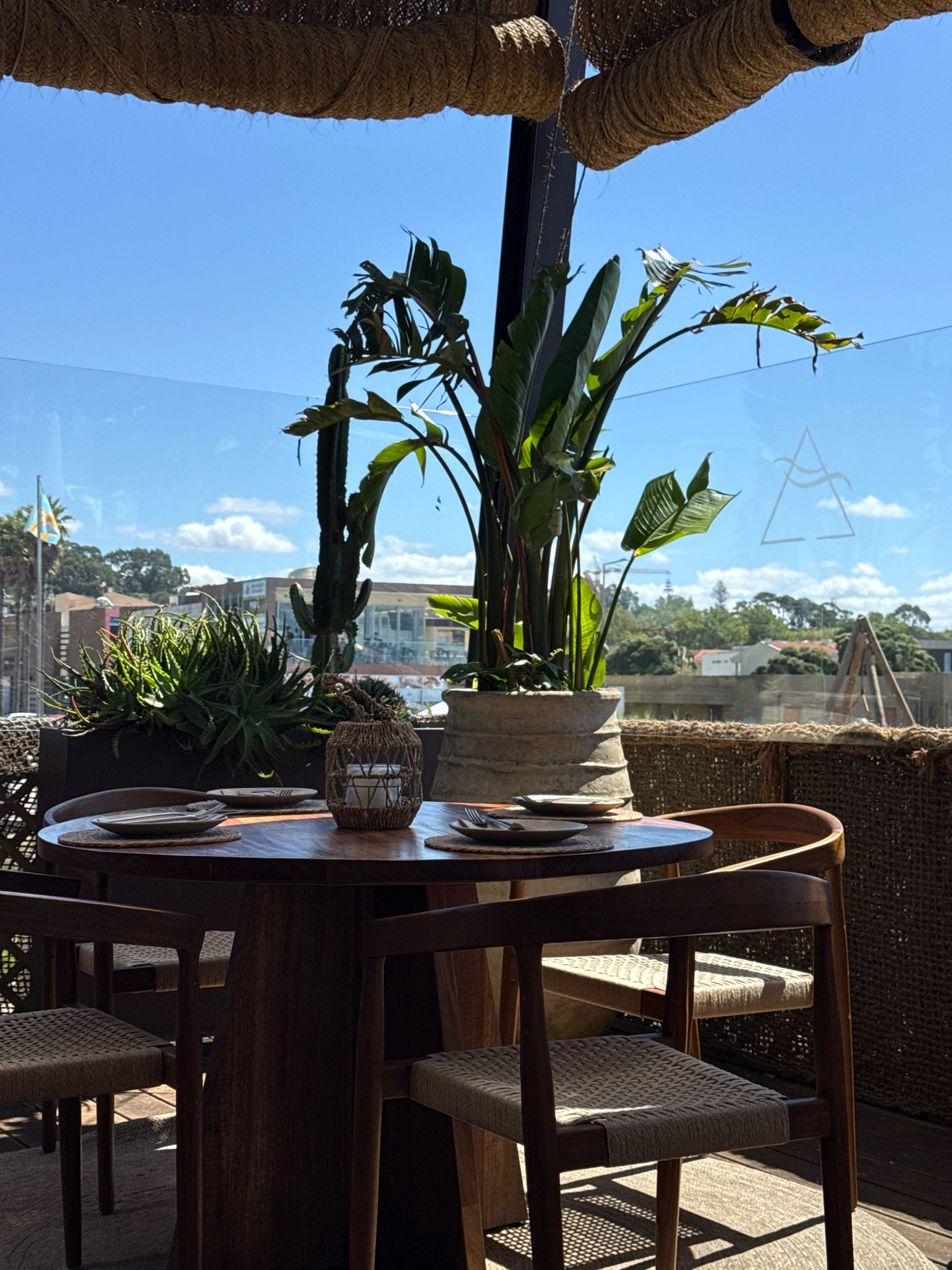 The image shows an outdoor seating area at a restaurant, possibly located in Vila Nova de Gaia, Portugal, with a view of the city skyline and the Douro River in the background. A round wooden table is set for dining with plates and cutlery. Surrounding the table are several chairs made of wood and woven material. In the foreground, a large potted plant with broad leaves adds a touch of greenery. In the background, a flag is visible on a pole, and various buildings and trees are scattered across the landscape. The sky is blue with scattered white clouds, and the sun is casting shadows on the ground, indicating it is daytime. A logo or symbol, appearing to be a stylized triangle with wavy lines inside, is visible on a glass barrier. The overall scene depicts a relaxed and inviting atmosphere for dining.