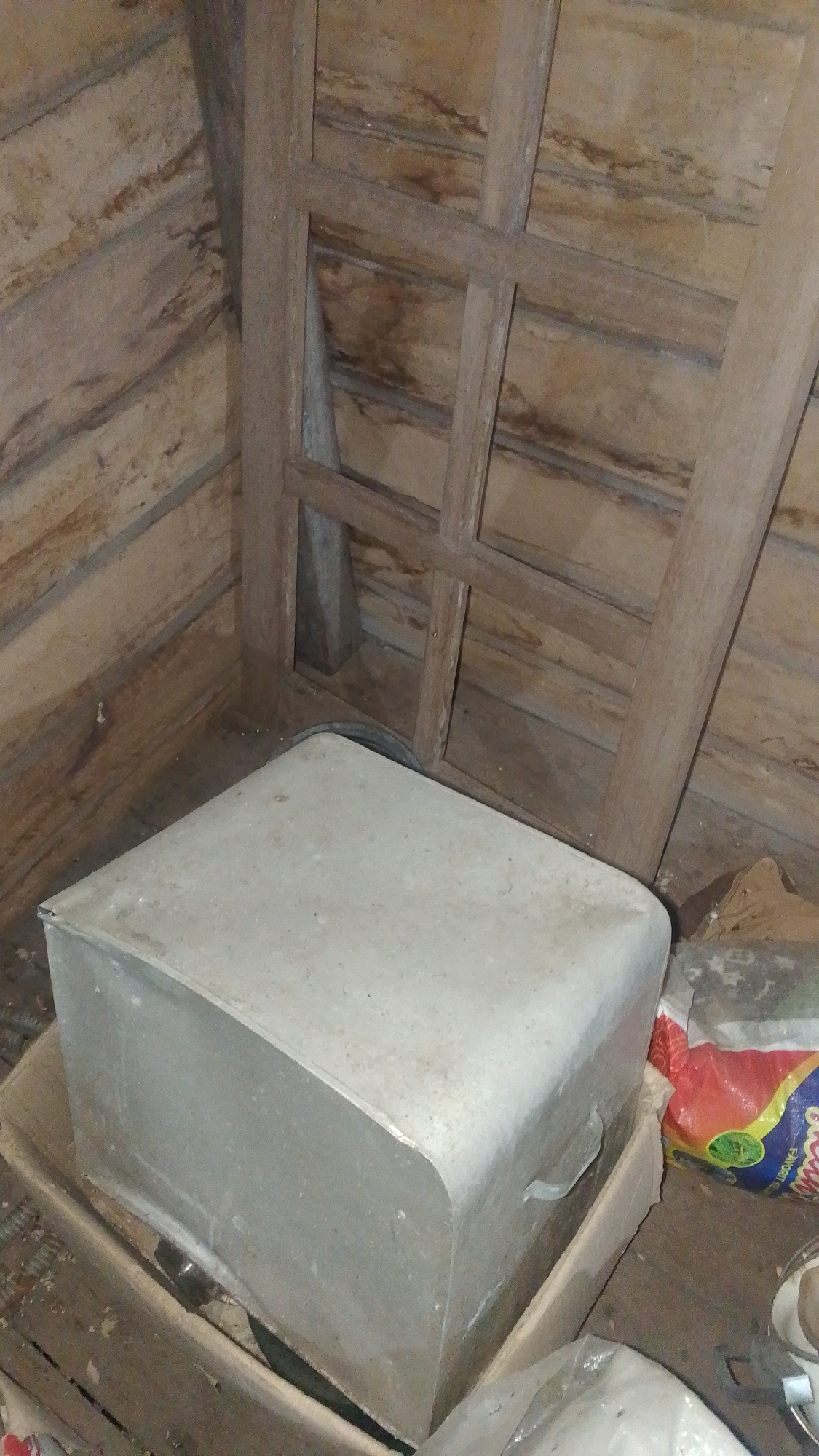 In a dimly lit, rustic wooden structure, likely an attic or storage space, a large, rectangular, light grey metal container rests on a wooden base. The container has rounded edges and what appears to be a handle on its right side. The interior walls of the structure are made of horizontal wooden planks, some showing signs of age and staining. A wooden frame, resembling part of a window or partition, stands vertically in the background. To the right of the metal container, a colorful plastic bag, possibly containing food items, is visible, along with other unidentifiable objects. The floor is also wooden. The scene is devoid of human presence. The overall impression is one of an unused, dusty storage area within a building. The location is identified as Olak-Olak Kubu, Indonesia.