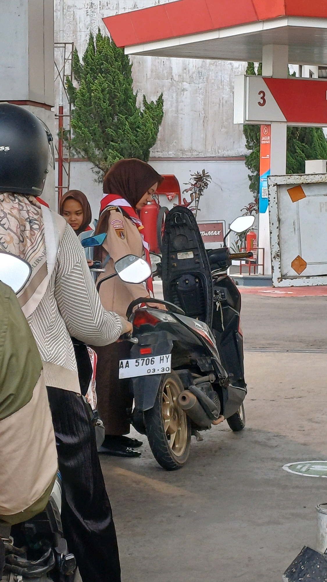 This is a medium shot of a gas station in Kranggan, Indonesia, with three individuals and two motorcycles. In the foreground, a person wearing a black helmet and a patterned scarf is partially visible, facing away from the camera. To their left, another individual wearing a brown hijab and a brown uniform with a red and white sash is looking down at their phone. Behind them and slightly to the left, a third person also wearing a brown hijab is visible. A black scooter is parked in the center of the frame, with its seat open. The gas station features red and white signage, with a "3" prominently displayed on one of the pump covers. Green cypress trees are visible behind the gas station. The ground is paved with concrete.