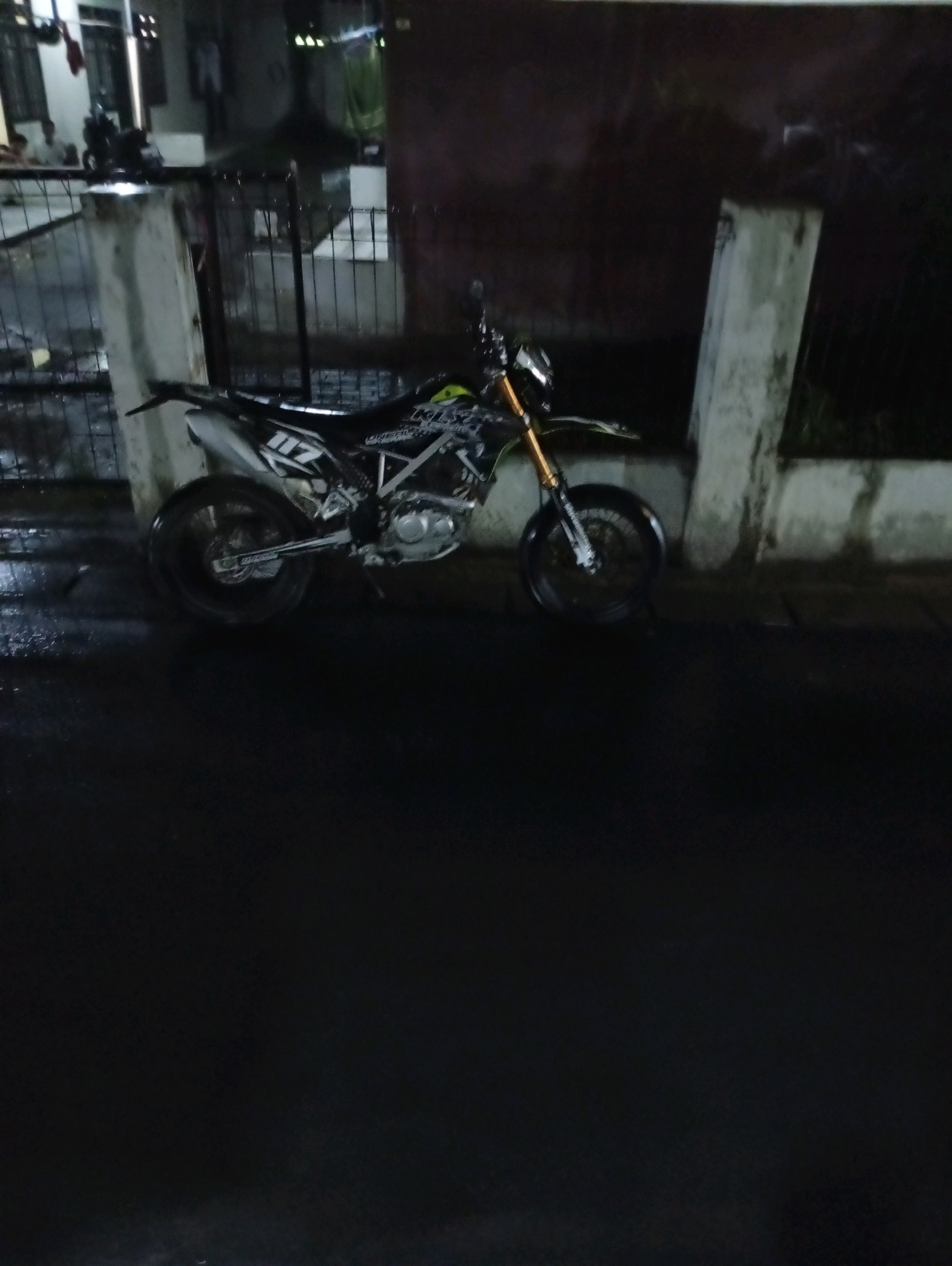 A dirt bike is parked on a wet street at night. The bike is black and white with yellow accents and the number 117 is visible on the side. A concrete pillar and fence are behind the bike, and a dark wall is in the background. The street is wet, reflecting the lights from the surrounding area. The scene appears to be in a residential or urban area, possibly in Karawang, Indonesia, given the context. There are no people clearly visible in the image.
