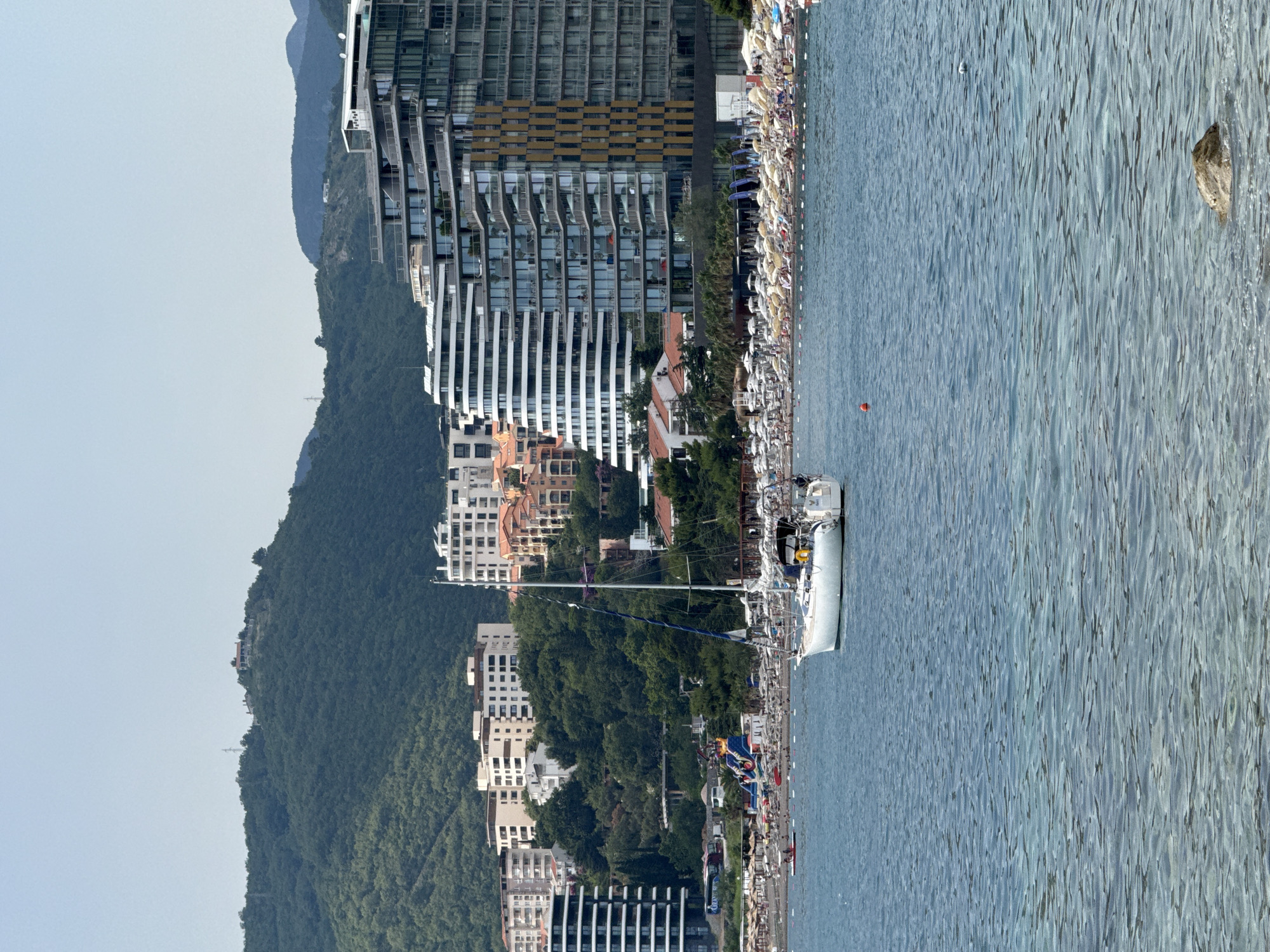 This media file depicts a vibrant coastal scene in Bečići, Montenegro. The image is oriented vertically, showcasing a densely populated beach with numerous sunbathers and beach umbrellas lining the shore. Adjacent to the beach is a modern, multi-story building with a striking architectural design featuring glass facades and horizontal elements. Further inland, lush green hills rise steeply, dotted with buildings and trees, suggesting a mountainous backdrop.
The sea is a prominent feature, with gentle ripples on its surface and a few boats anchored near the shore. A white sailboat is visible docked. The presence of people on the beach and the clear sky suggest a pleasant day, likely in the afternoon, with fair weather suitable for outdoor activities. The combination of modern architecture, natural landscape, and beach activity points to a popular tourist destination. There are no visible text elements in the image.