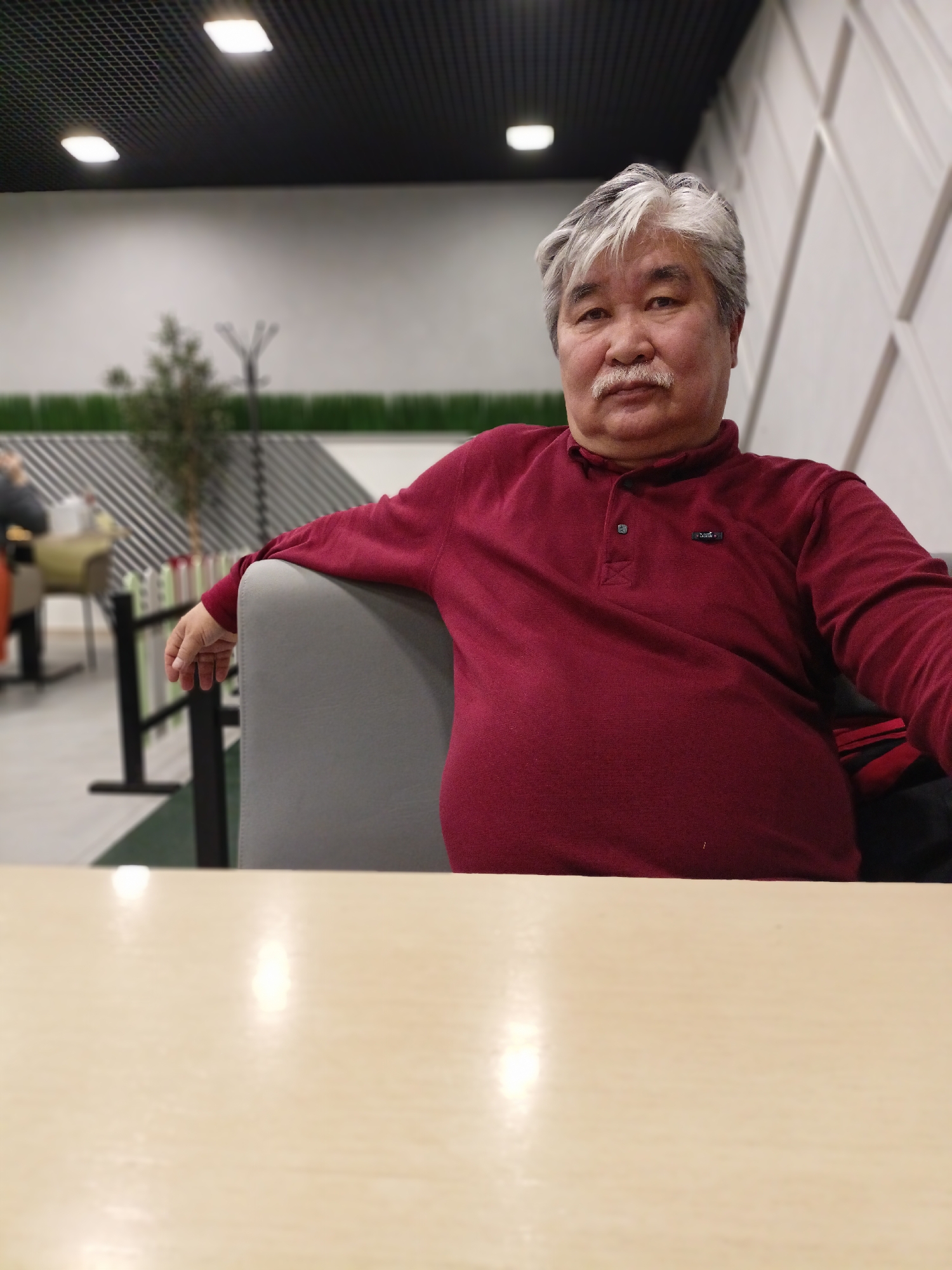 An Asian man with graying hair and a mustache is seated in a chair at a table. He is wearing a maroon polo shirt. He is in a room with a black grid ceiling and off-white walls with geometric paneling. A plant and coat rack are visible in the background. The location is Novosibirsk, Russia.