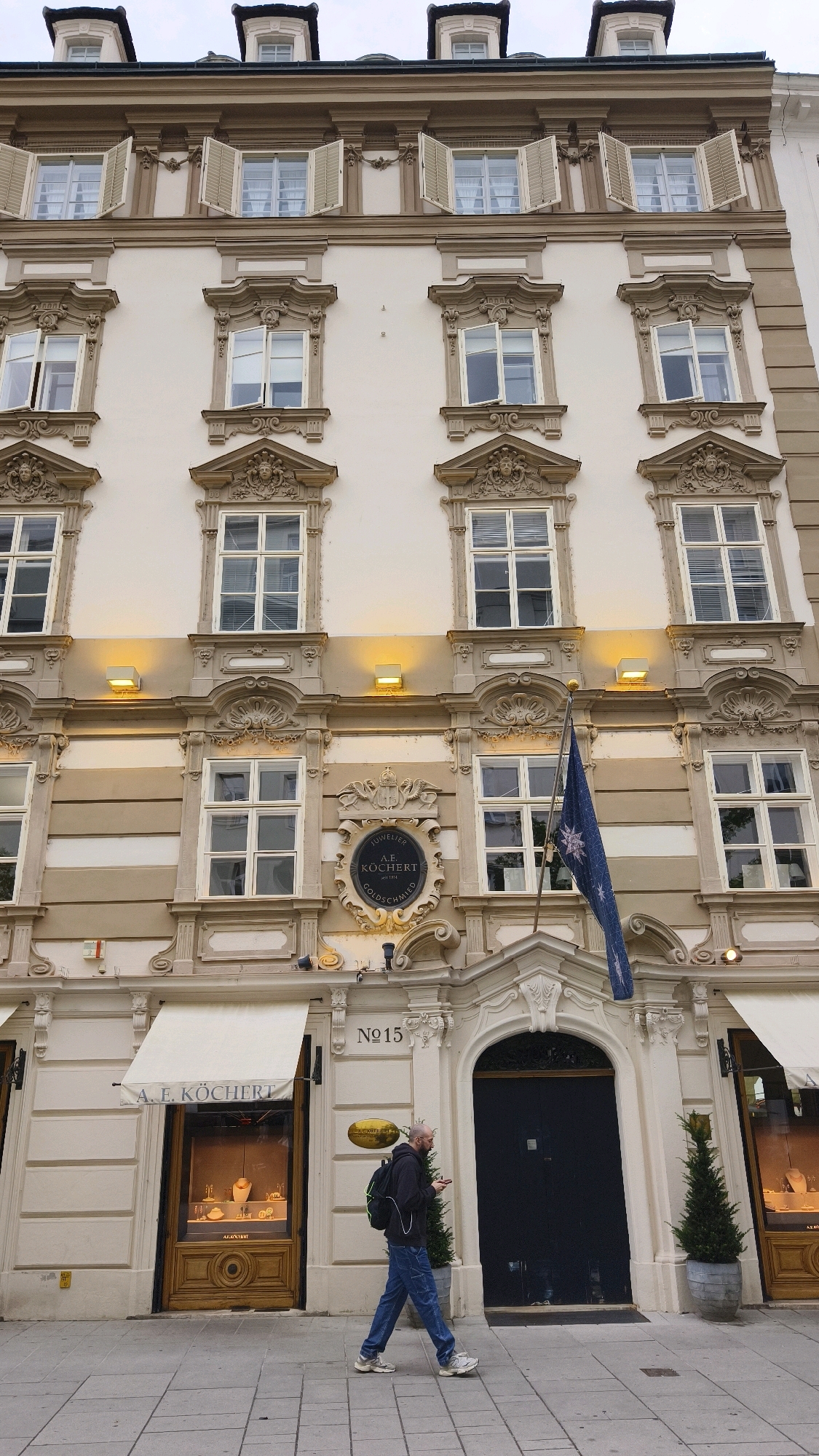 The image shows the facade of a multi-story building in Vienna, Austria. The building is ornately decorated with architectural details, including multiple windows with shutters and elaborate framing. A blue flag with a white star pattern is hanging from a flagpole in front of the building. The ground floor features a shop with a large display window showcasing jewelry. A man wearing a backpack and jeans is walking in front of the shop. Above the shop entrance, the number "15" is visible, along with a circular emblem that reads "JUWELIER A.E. KÖCHERT GOLDSCHMIED". To the right of the shop entrance, a large dark door is visible, and to the right of that, another shop window. The street is paved with cobblestones. The sky is overcast.