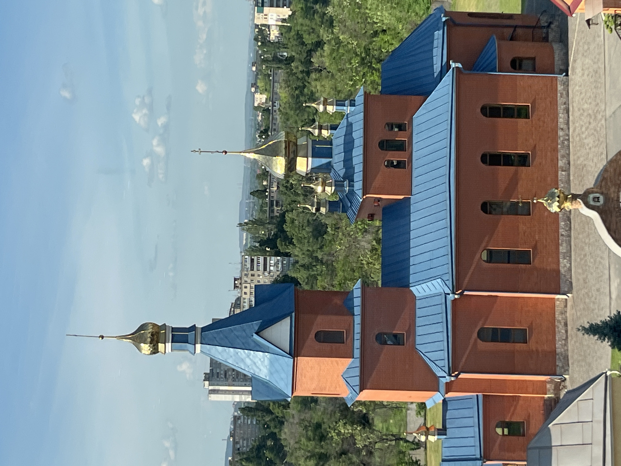 The image depicts an elevated, oblique view of a religious building, likely an Orthodox church, situated in the city of Kryvyi Rih, Ukraine. The architecture is characterized by a brick facade and a blue metal roof. The building features distinct golden domes topped with crosses, common in Orthodox Christian architecture. The sky is visible in the background, exhibiting a clear, blue color with sparse cloud cover. Surrounding the church are green trees and urban structures, suggesting its location within a developed part of the city. The image appears to be taken from a high vantage point, possibly from an adjacent building or an elevated structure.