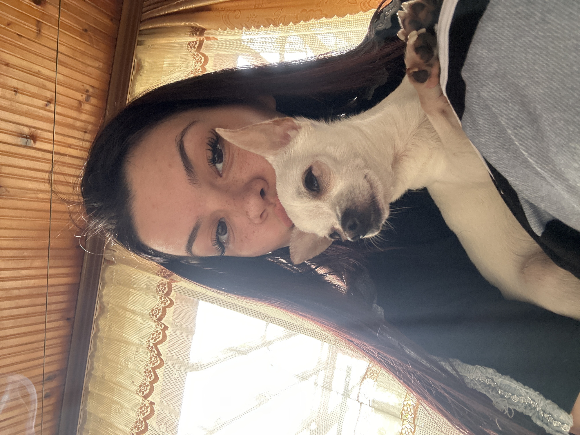 A woman with long dark hair and freckles is holding a small, white dog. They appear to be indoors, with wood paneling on the wall and lace curtains visible behind them. The dog's paws are visible on the woman's shoulder.
The location is not specified in the picture.