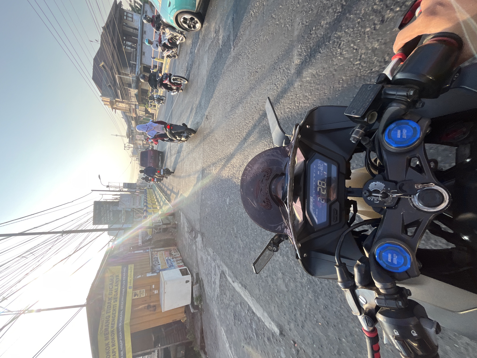 The image captures a first-person perspective of riding a motorcycle on a street in Kalinyamatan, Indonesia, during sunset. The road is asphalt, showing signs of wear and tear, with some potholes. Various vehicles, including motorcycles, cars, and a van, are on the road, suggesting moderate traffic. People on motorcycles are dressed in everyday attire, with some wearing helmets and headscarves. Buildings line the street, displaying a mix of commercial establishments, including a "CIGARETTE STORE" with a yellow banner, and residential structures. Overhead, a dense network of power lines crisscrosses the sky. The sun is low on the horizon, casting long shadows and creating a bright glare that illuminates the scene. The motorcycle's dashboard displays the time as 2:28.