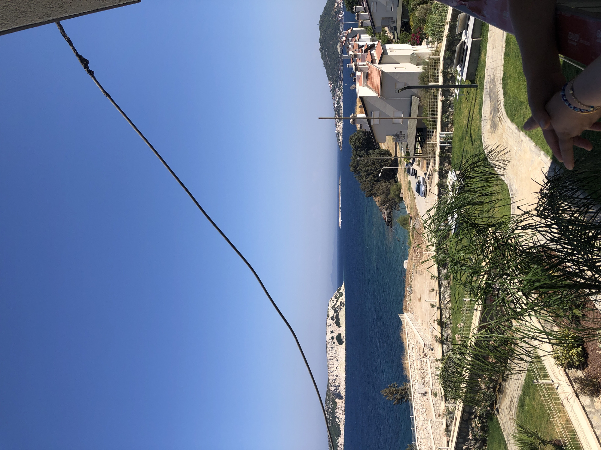 The image depicts a coastal view in Karaburun, Turkey, on a clear, sunny day. In the foreground, a person's hands are visible, resting on a railing. Beyond the railing, a well-maintained garden area features green grass, paved pathways, and lush vegetation, including palm fronds.
The middle ground showcases a row of white houses with red-tiled roofs, situated on a hillside overlooking the sea. A road with parked cars runs alongside the houses. The sea itself is a deep blue, stretching out towards the horizon.
In the background, a large, distinctive island with a flat top and sheer white cliffs dominates the left side of the frame. The island has patches of greenery and appears to have some natural rock formations or caves. Further in the distance, another, smaller hilly island with scattered buildings can be seen. A dark cable runs diagonally across the upper portion of the image, from the top right corner towards the center. The sky is a clear, bright blue.