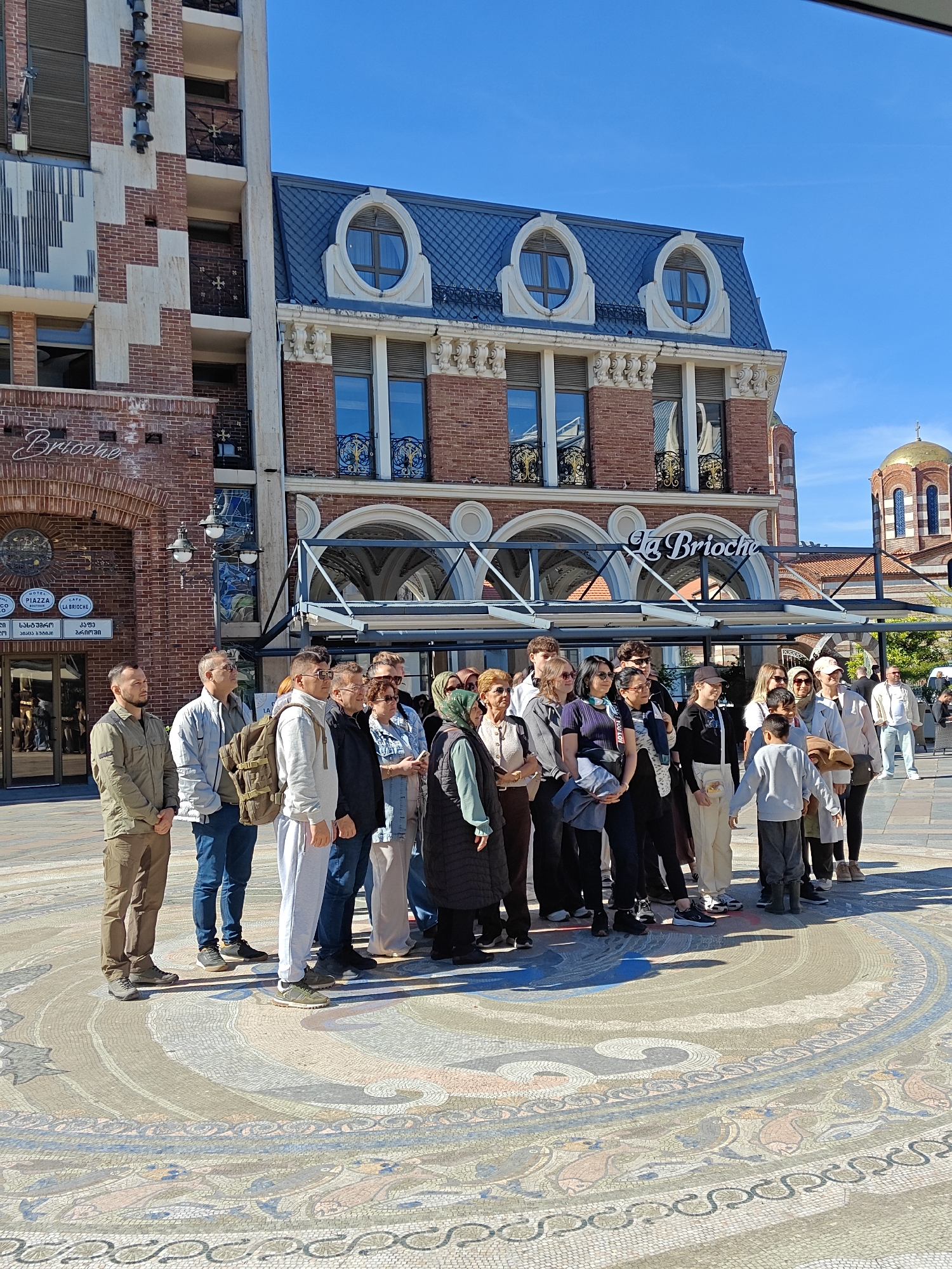 The image depicts a group of people standing on a mosaic-paved area in Batumi, Georgia. In the background are two brick buildings, one with the name "La Brioche" displayed on it. A church with a gold dome is visible to the right. The group of people is diverse in age and attire. The sky is clear and blue.