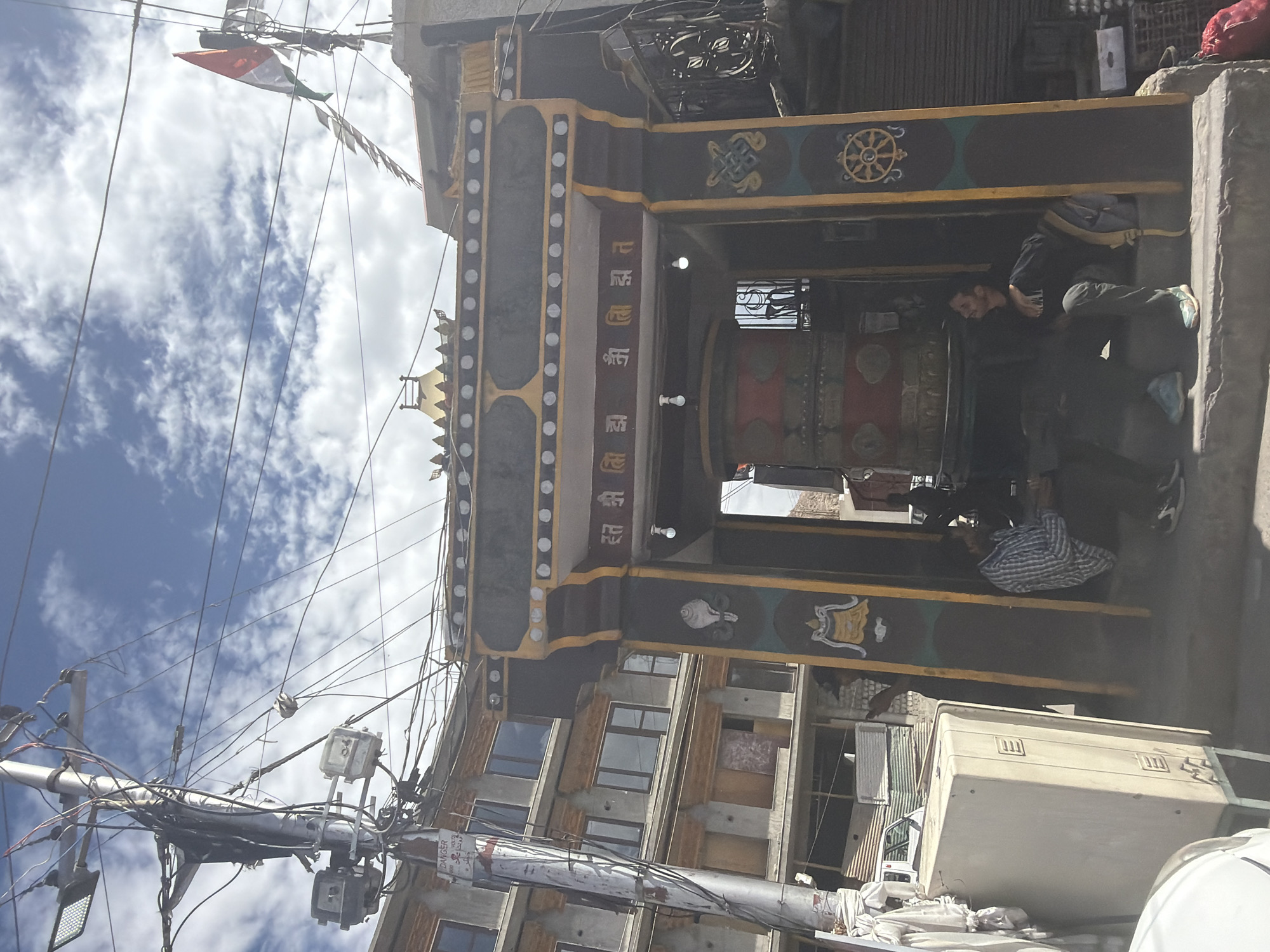 In Leh, India, a street scene unfolds under a cloudy sky. A prominent electrical pole with a dense tangle of wires stands to the left, adorned with multiple light fixtures and a "DANGER HIGH VOLTAGE" sign. Adjacent to it, a multi-story building with grey facades and tinted windows is visible. In the foreground, a large, cream-colored rectangular object, possibly a storage unit or utility box, rests on a metal frame.
Dominating the center is a decorative archway, characteristic of Tibetan Buddhist architecture, featuring intricate patterns in yellow, maroon, and black. Inscribed above the archway are Tibetan characters. Within the archway, a large, red and gold prayer wheel is the central focus. Two individuals are seated on the steps leading up to the archway. One person, wearing a blue and white checkered shirt, is looking away from the camera. The other, in a dark shirt and grey pants, is looking upwards. A small Indian flag with red, white, and green colors is visible on a pole on top of a structure in the background.