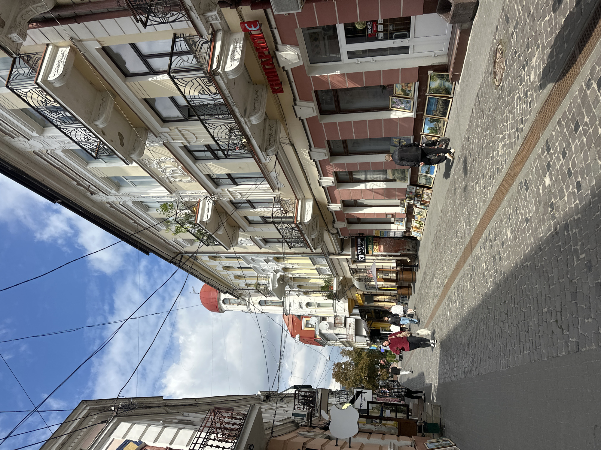 The image captures a street scene in Ternopil, Ukraine. The street is paved with cobblestones and lined with buildings featuring balconies with ornate metal railings. In the foreground, a man in a black jacket walks away from the camera, passing a display of framed paintings and artwork laid out on the ground in front of a pink-colored building. Further down the street, several people are walking. The buildings showcase architectural details, with ornate moldings and varying colors. A prominent red-domed tower is visible in the background. Overhead, a network of electrical wires crisscrosses the sky, which is partly cloudy. Signs in Cyrillic script are visible on some of the buildings, indicating shops or businesses. The overall impression is of a historic urban street with pedestrian activity and commerce.