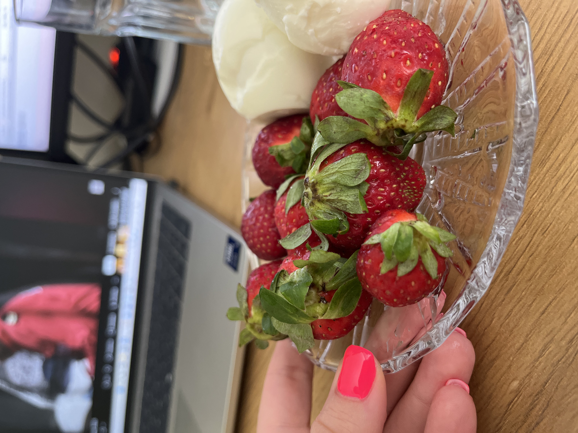In Plovdiv, Bulgaria, on a wooden table, a clear glass dish holds several ripe strawberries and two portions of white cheese. A person's left hand, with bright pink nail polish, is visible near the dish. In the background, a laptop is open with a blurred image on its screen, and a glass of water stands to the right. The scene appears to be a casual snack or meal setting.