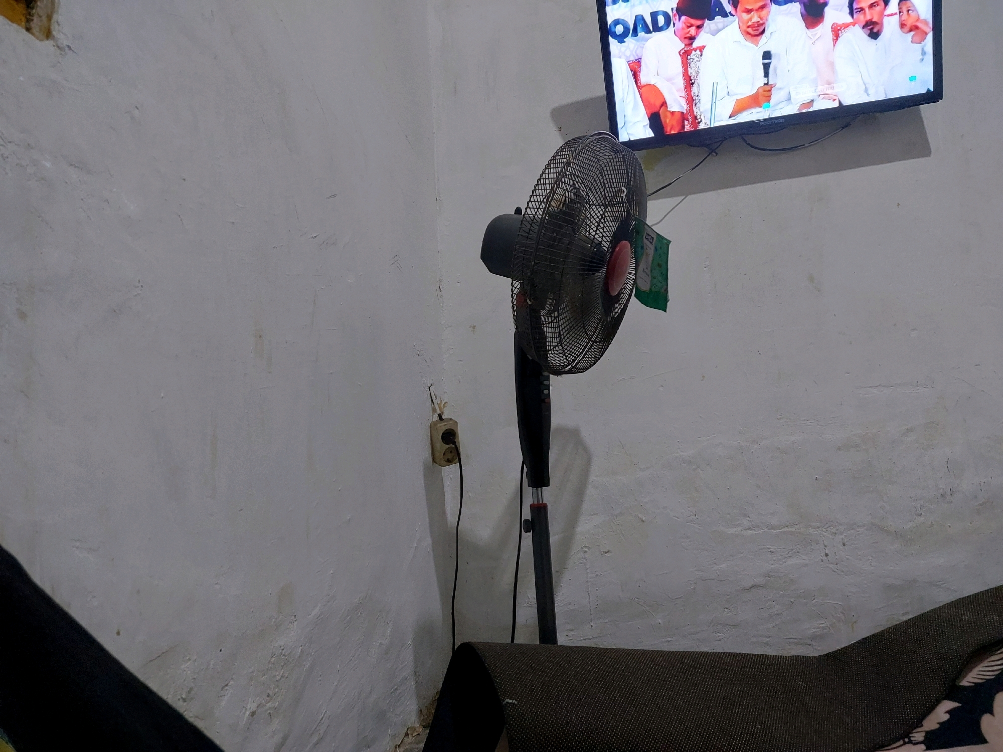 The image shows a dimly lit room with white walls. In the foreground, a black oscillating fan stands on a floor or low surface, with its grille visible and a green flag attached to it. Behind the fan, a black pole rises vertically, connected to the fan head. A power outlet with a black cord plugged into it is mounted on the wall to the left of the fan. Above the fan, a television screen is mounted on the wall, displaying an image of several men in white attire, some holding microphones. The room appears to be a residential or public space, possibly in Sarangan, Indonesia, given the context provided. The overall impression is one of a simple, perhaps humble, indoor environment.