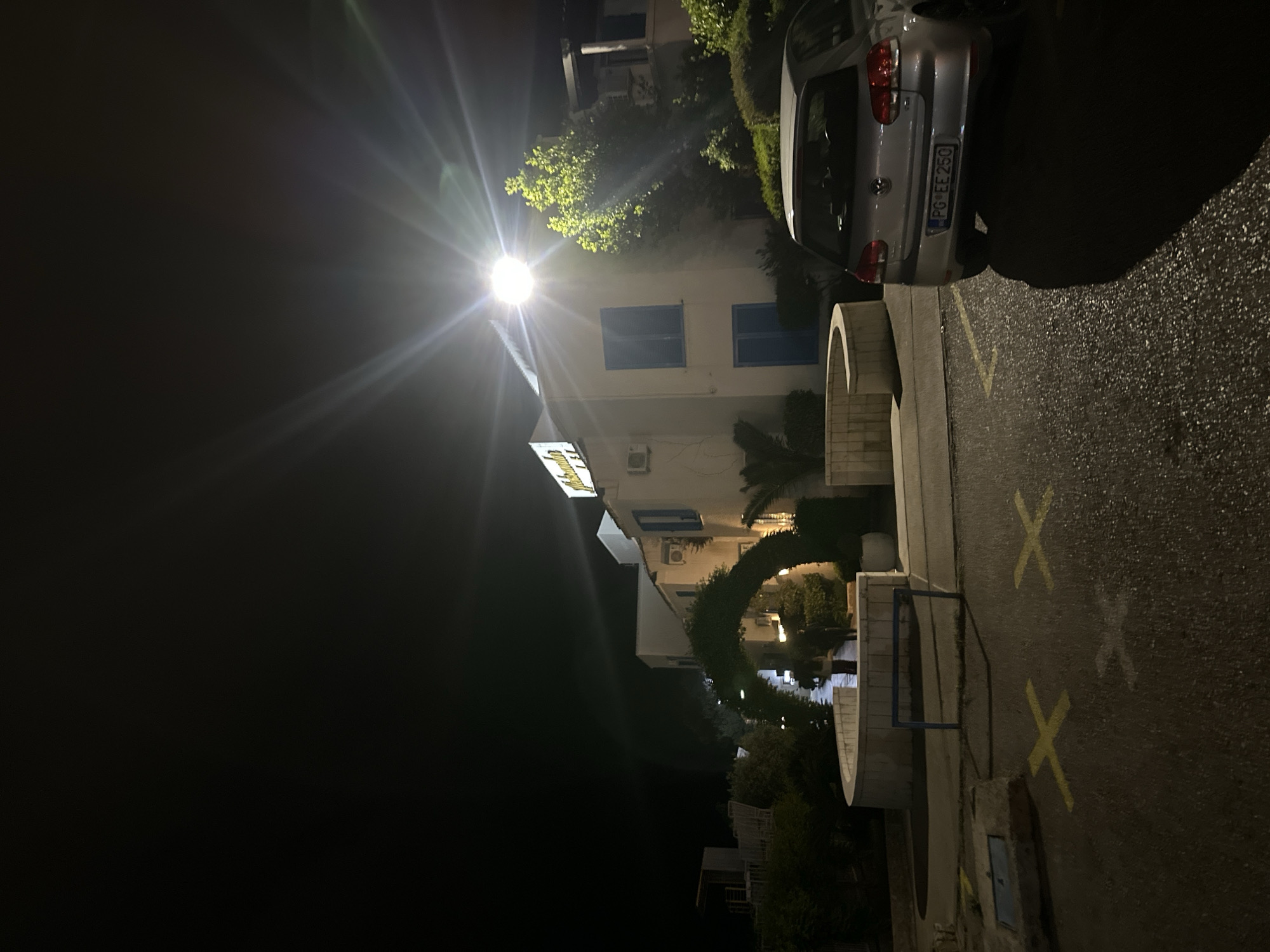 A nighttime view in Budva, Montenegro, shows a street lit by a bright street lamp, causing glare. A silver car, license plate PG EE 250, is parked on the right side of the road. The road surface is dark and marked with yellow painted X's. To the left of the car is a white building with blue window shutters. The building's name is illuminated above the entrance. Plants surround the building and an arched plant tunnel is visible in front of the building. The sky is dark.