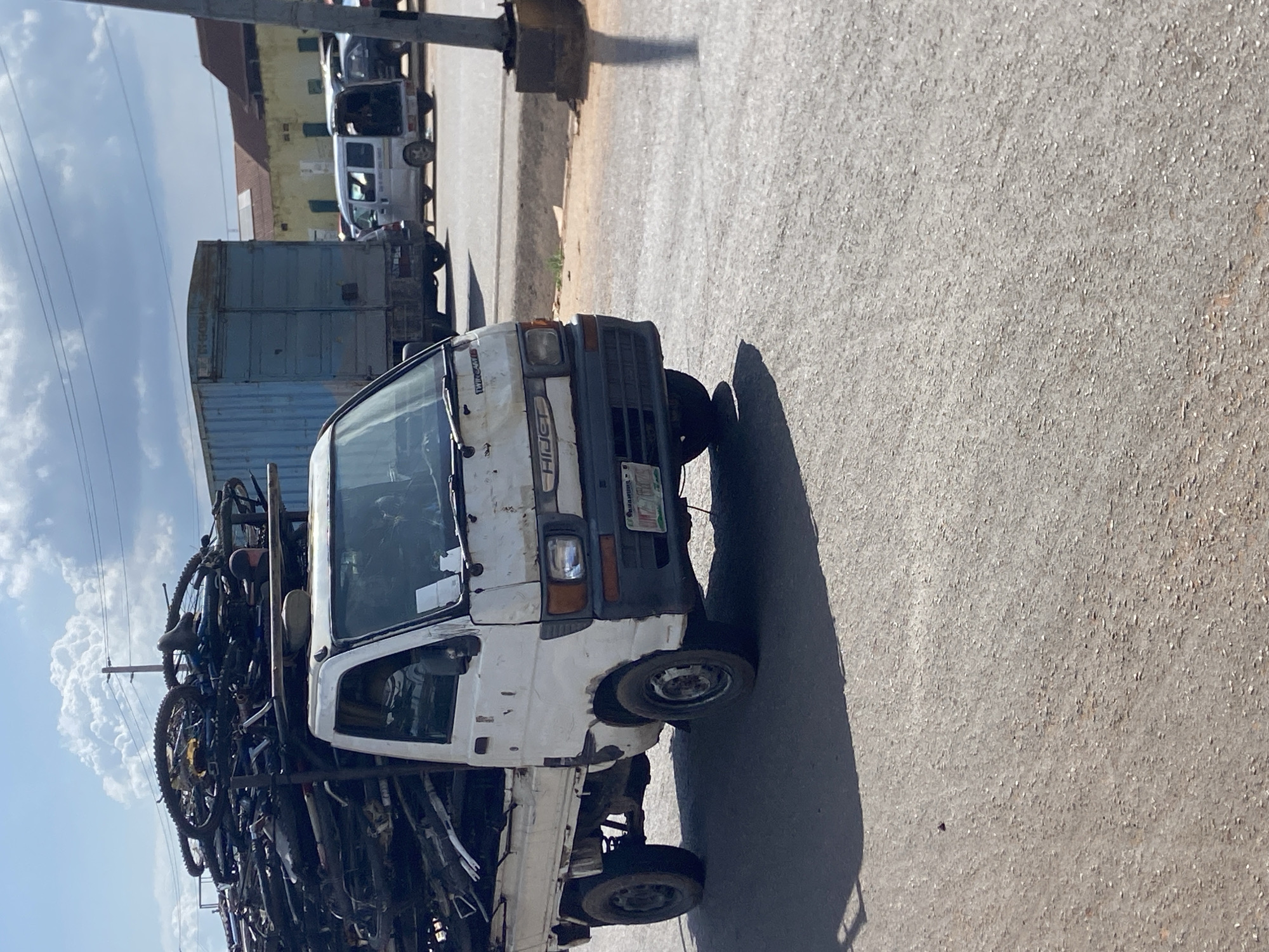 A white Daihatsu Hijet truck is parked on an asphalt road in Ogidi, Nigeria, under a partly cloudy sky. The truck bed is overloaded with a large pile of bicycles, most of which appear to be used or dismantled. The truck has a gray bumper and a dented front fender. The license plate is visible, displaying green lettering on a white background. In the background, a large blue shipping container truck is parked on the right, with a smaller white van to its right. Buildings with red roofs and yellow walls are visible behind the vehicles, suggesting an urban or semi-urban setting. The ground is predominantly asphalt, with some patches of dirt and sparse vegetation visible near the road's edge. The scene appears to be a roadside area, possibly a marketplace or a loading zone.