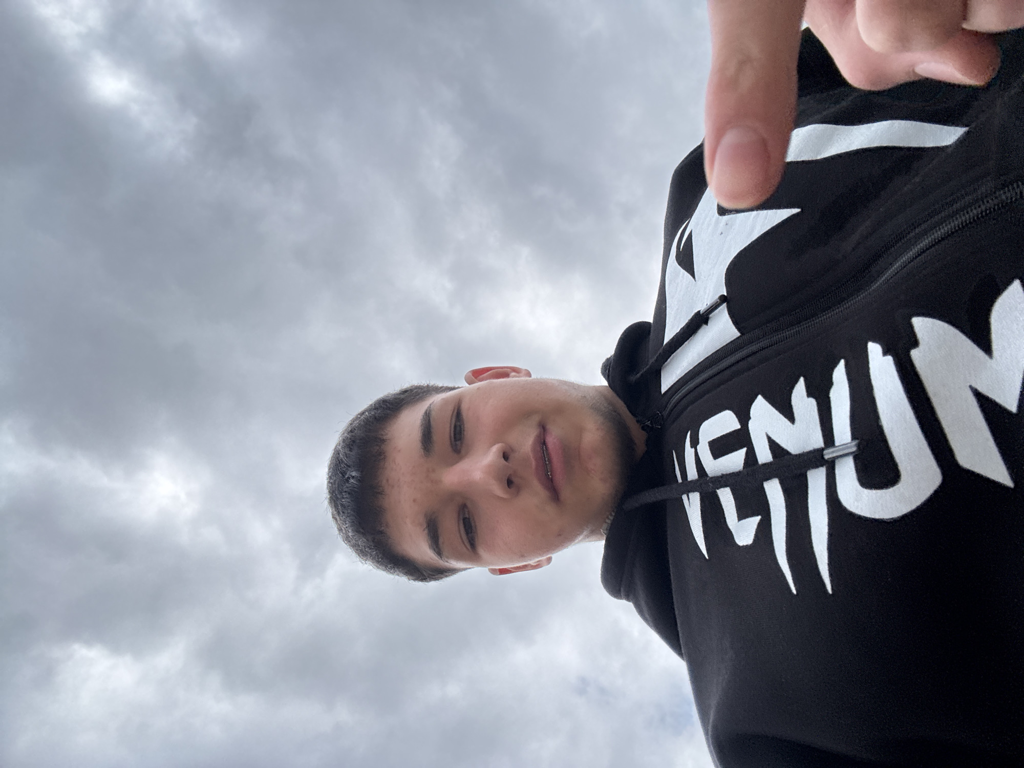 A young man with short dark hair and a slight beard is looking down at the camera. He is wearing a black hoodie with white lettering. Part of his thumb is visible at the top of the frame. The background is a cloudy sky. The location is unknown, and there is no evidence to suggest it is Zlatograd, Bulgaria.