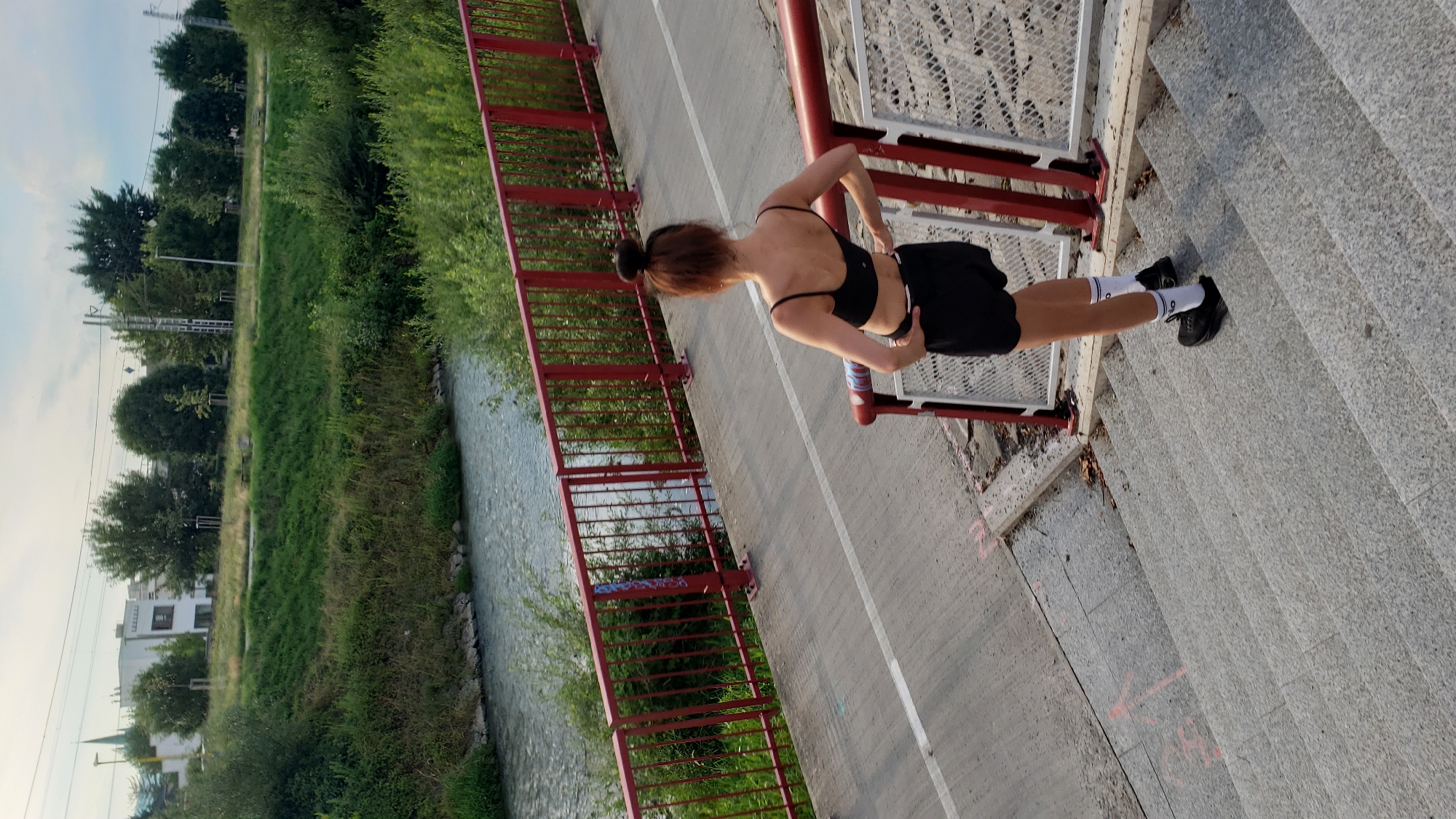 A full, outdoor, eye-level shot shows a woman with her back to the camera, standing on a concrete staircase next to a red metal bridge railing. She is wearing a black sports bra, black shorts, white socks, and black sneakers. Her hair is pulled up into a bun on top of her head. The bridge spans a body of water that is bordered by a grassy slope and trees. In the distance, a white building with a pointed spire is visible, along with more trees and a utility pole. The sky is overcast. The image appears to be taken in the city of Prešov, Slovakia, possibly near the Torysa river.