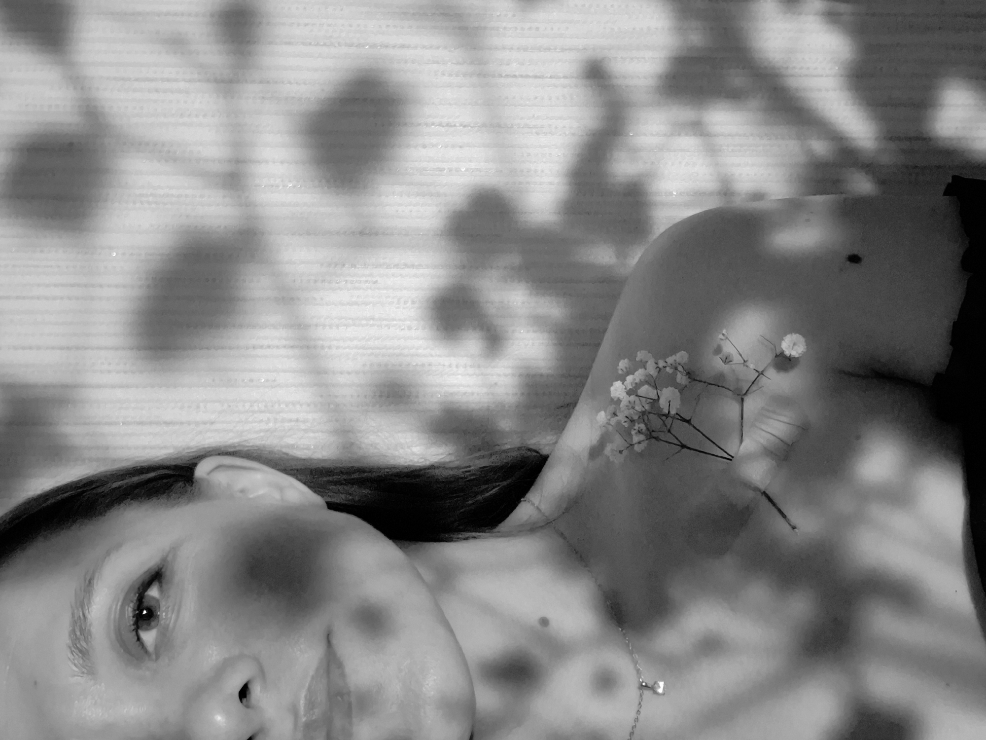 A black and white photograph shows a woman lying down, presumably indoors. Her head is tilted, with her face partially obscured by shadows. She is wearing a necklace with a small pendant. On her right shoulder, there is a sprig of small white flowers. The background is a wall covered in shadows that resemble foliage. The location of Chaikovsky, Russia, is not determinable from the image.