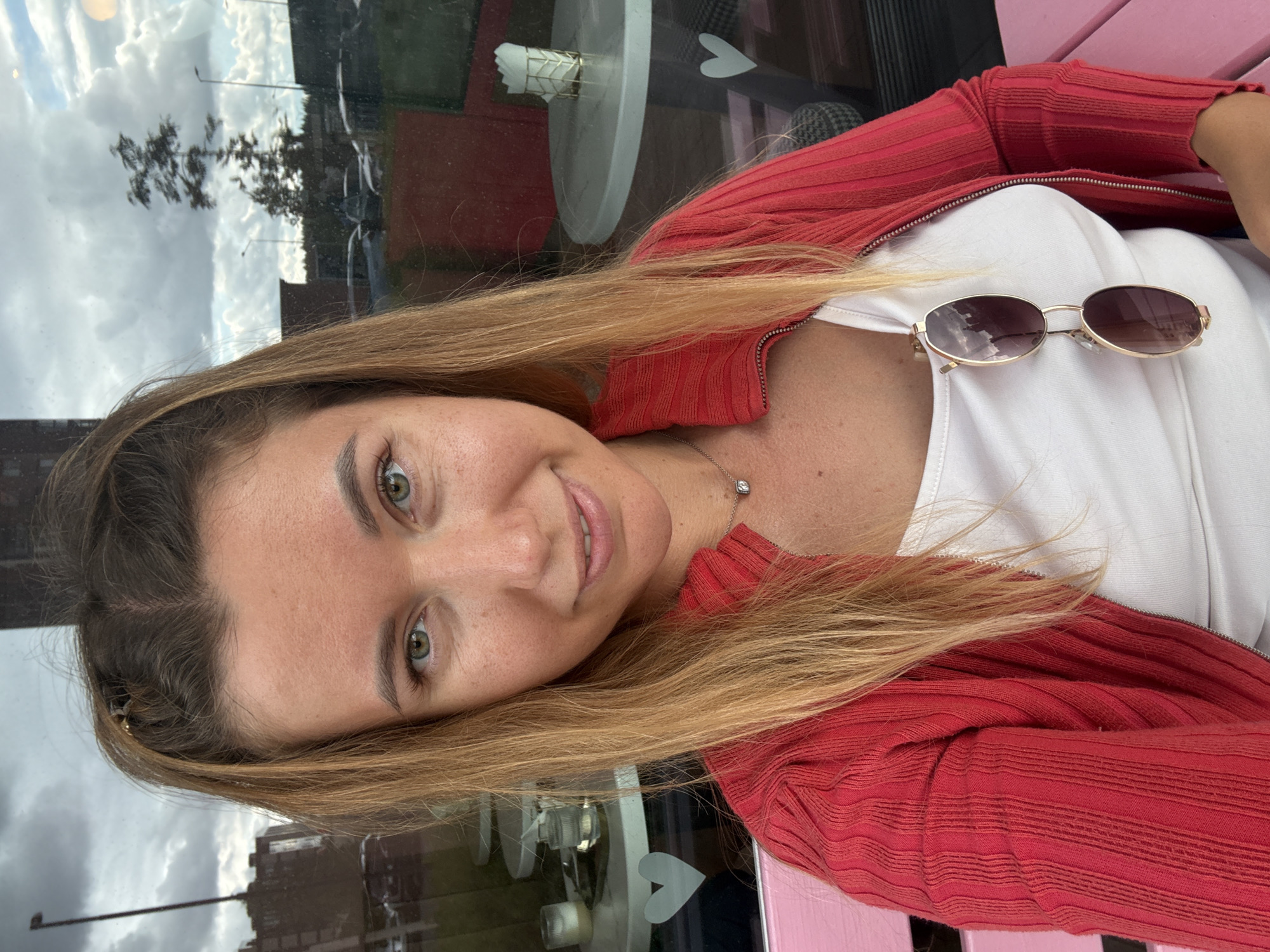 A young woman with blonde hair pulled into two small buns on the top of her head, with the rest of her hair down, is pictured in a selfie. She is wearing a red zip-up cardigan over a white tank top, and a thin silver necklace with a diamond pendant. Sunglasses are clipped to the front of her tank top. She is sitting on a pink bench outside a building with large windows, which reflect a cloudy sky, trees, cars, and streetlights. A small table with a candle and a napkin holder can be seen through the window behind her.
