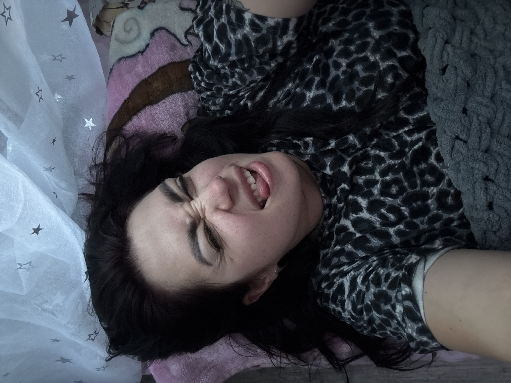 A woman is lying down, possibly in bed, with her eyes squinted and her tongue sticking out. She is wearing a leopard-print shirt and is covered by a gray blanket. A white sheer fabric with stars printed on it is draped above her. The background appears to be a bed with a pink and brown patterned blanket.