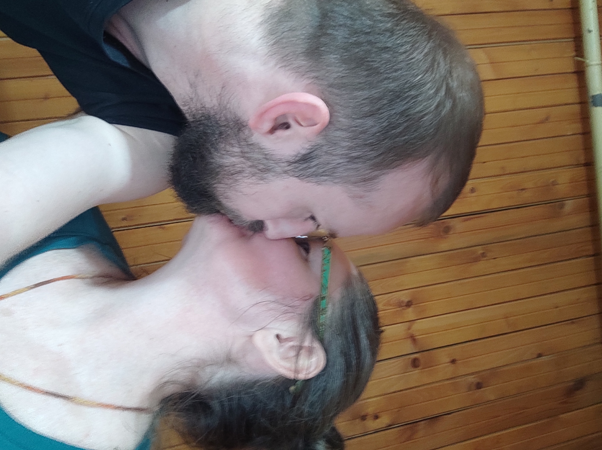 A couple is kissing in front of a wooden wall. The man has short brown hair, a beard, and is wearing a black shirt. The woman has grey hair pulled back, a gold necklace, and is wearing a teal shirt. The location is not discernible, nor can it be determined whether the scene is in Kratovo, Russia.