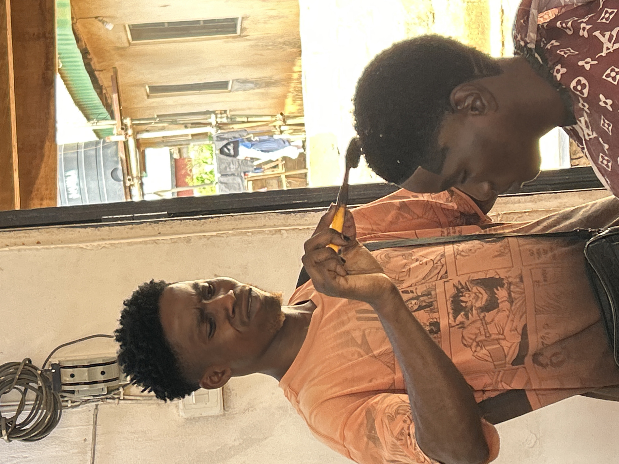 Two men are visible in the scene. One man is holding a small, yellow-handled object. The location appears to be outdoors, with a building and other structures visible in the background. The exact city and further contextual details are indeterminate from the provided image.