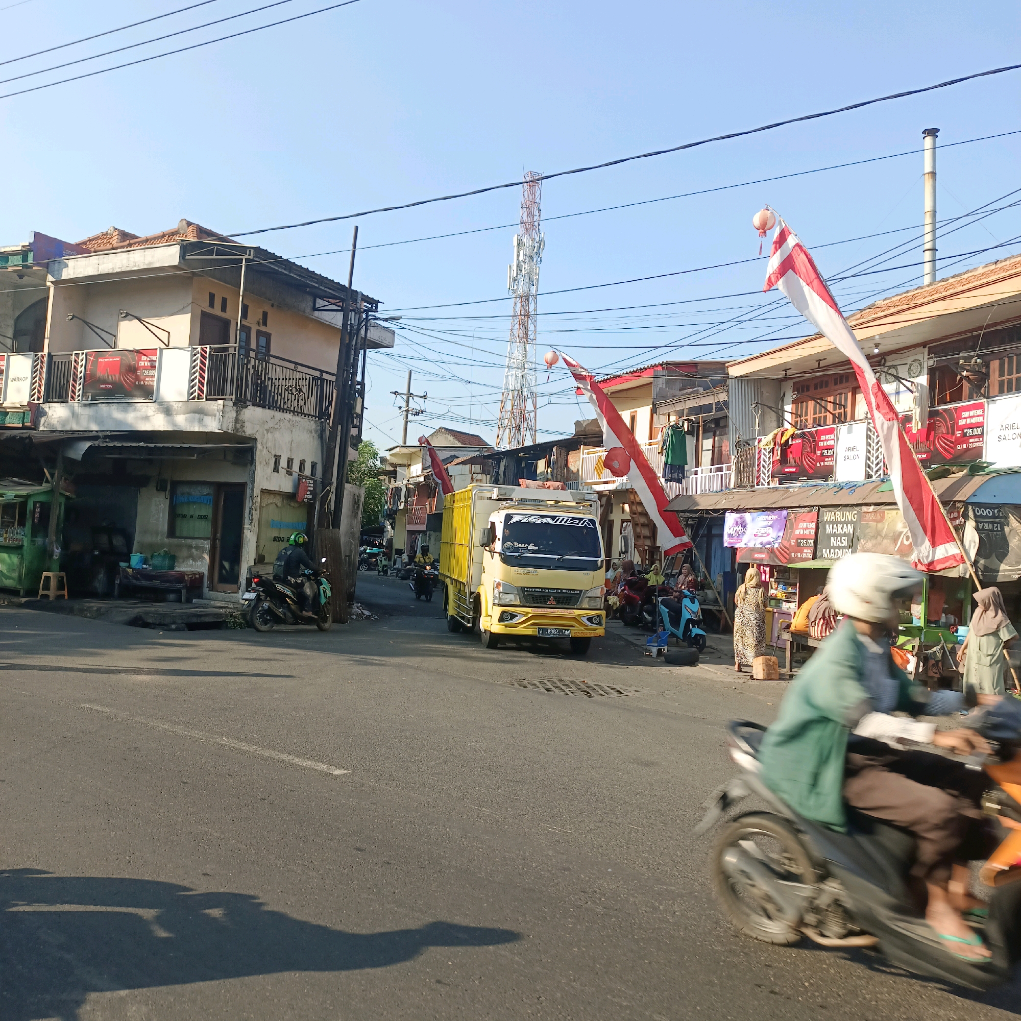 The image depicts a street scene in Surabaya, Indonesia, likely during daylight given the bright sun and clear sky. The street is paved and appears to be a local road with shops lining both sides. In the foreground, a person on a motorcycle is captured in motion, their form blurred due to the camera's focus and movement. The rider wears a helmet and a light green long-sleeved shirt. Behind them, other motorcycles and some people are visible, suggesting a moderately busy commercial area.
A yellow truck with "Bismillah" written on its windshield is prominently positioned in the middle of the road, moving away from the viewer. The truck has a flatbed with some cargo covered by a tarp. Various shops are visible, some with awnings and signage. One building on the left has a second story with a balcony and what appears to be an advertisement for "WARKOP" (a type of Indonesian coffee shop) and "STAY INTENSE 25.000". Indonesian flags, red and white, are displayed on poles along the street, indicating a possible celebration or national event.
In the background, a tall telecommunications tower stands against the clear blue sky, surrounded by numerous power lines. The architecture of the buildings suggests a mix of older and newer structures, common in urban Indonesian settings. There are also several small food stalls or kiosks. The overall atmosphere appears to be that of a typical bustling street in a provincial city, with everyday commerce and transportation activities in progress.
