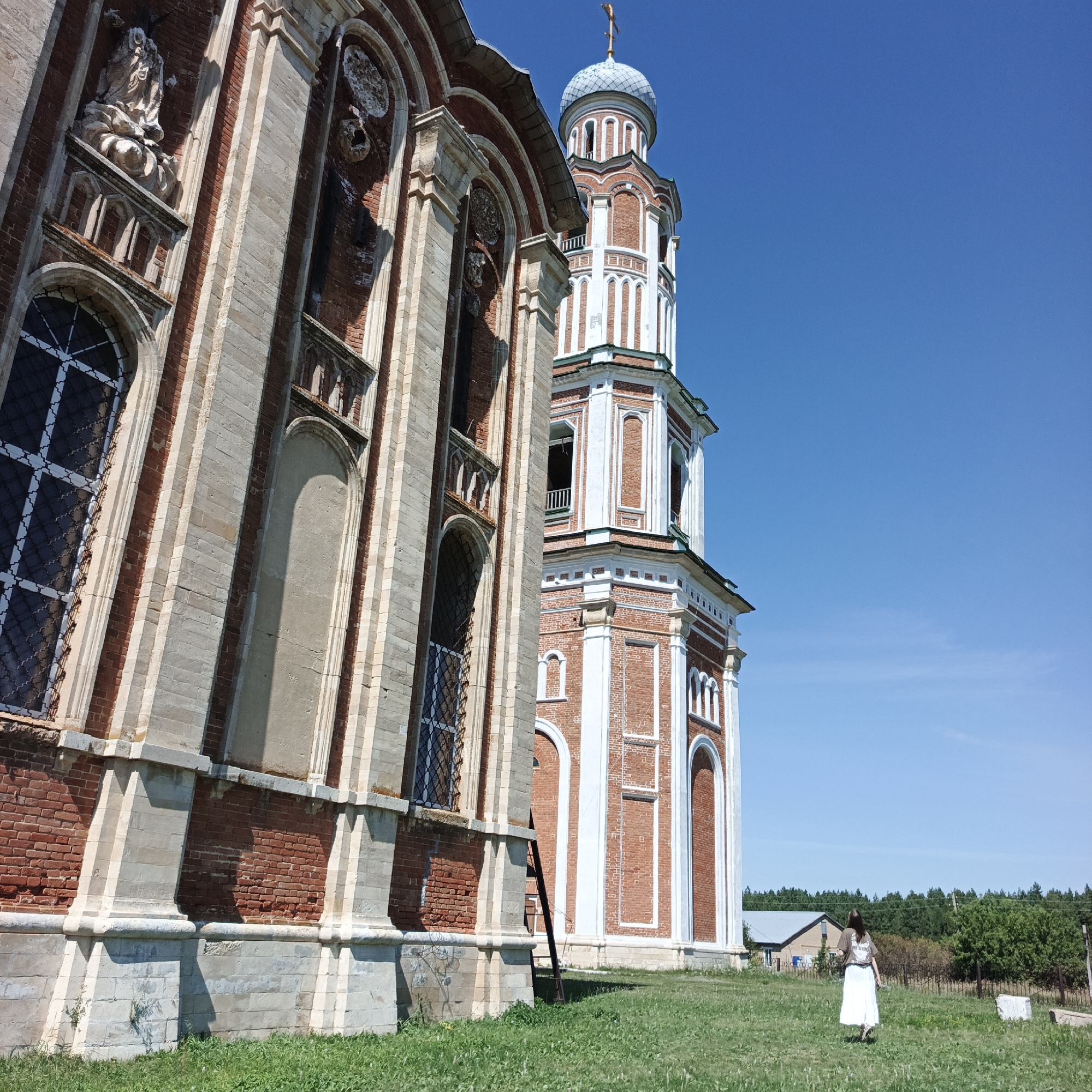 The image depicts the Church of the Assumption of the Blessed Virgin Mary in the village of Tugustemir, Russia, under a clear blue sky. The church's facade, constructed of red brick and adorned with white decorative elements, dominates the left side of the frame, showcasing intricate architectural details. A tall bell tower with a domed roof and a cross at its apex rises on the right. In the middle ground, a woman in a white skirt and a dark t-shirt walks away from the camera across a grassy field. In the background, a treeline and a building with a grey roof are visible. The overall scene suggests a bright, sunny day.