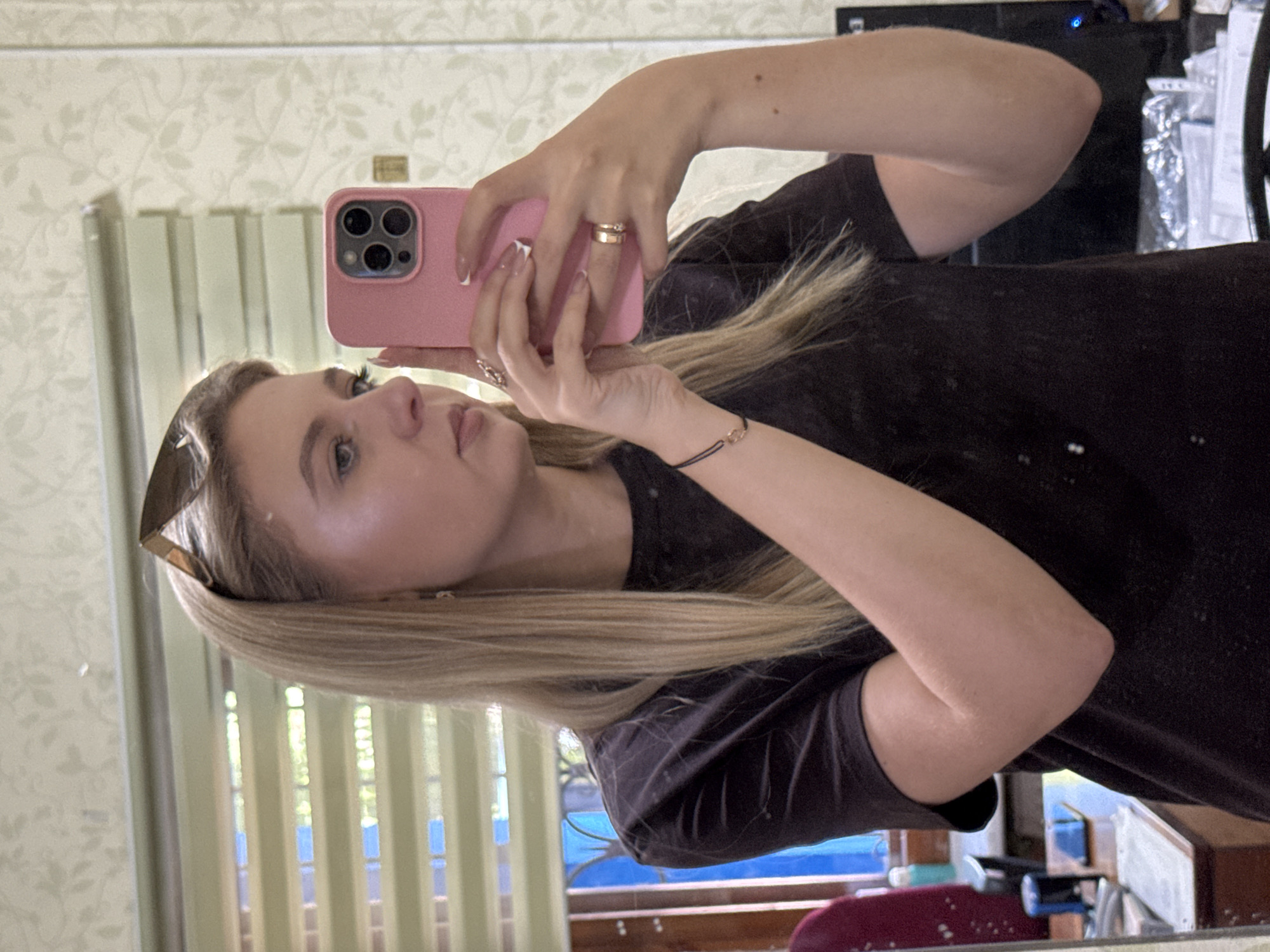 A young woman with blonde hair, wearing sunglasses on her head and a dark t-shirt, is taking a selfie in a mirror. She is holding a pink smartphone in her right hand, which is reflected in the mirror along with her face and upper body. She is wearing jewelry on her left hand, including a thin bracelet with a small gold circle and two rings. The background appears to be a room with blinds on a window, patterned wallpaper, and some furniture and objects, including a monitor, in the reflection.